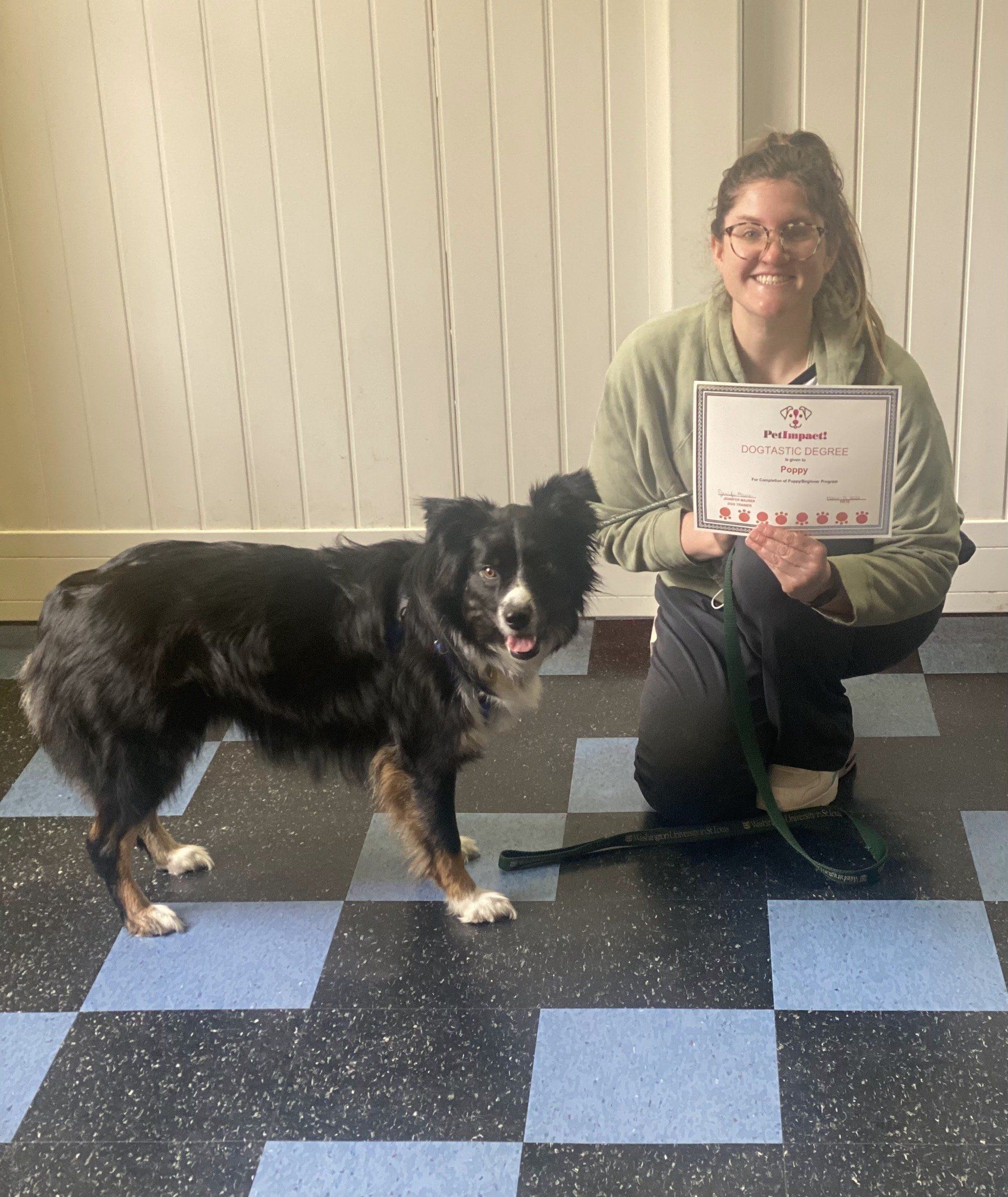 A woman kneels beside a dog, holding a certificate. They are on a checkered floor. The dog is black, gray, and brown.