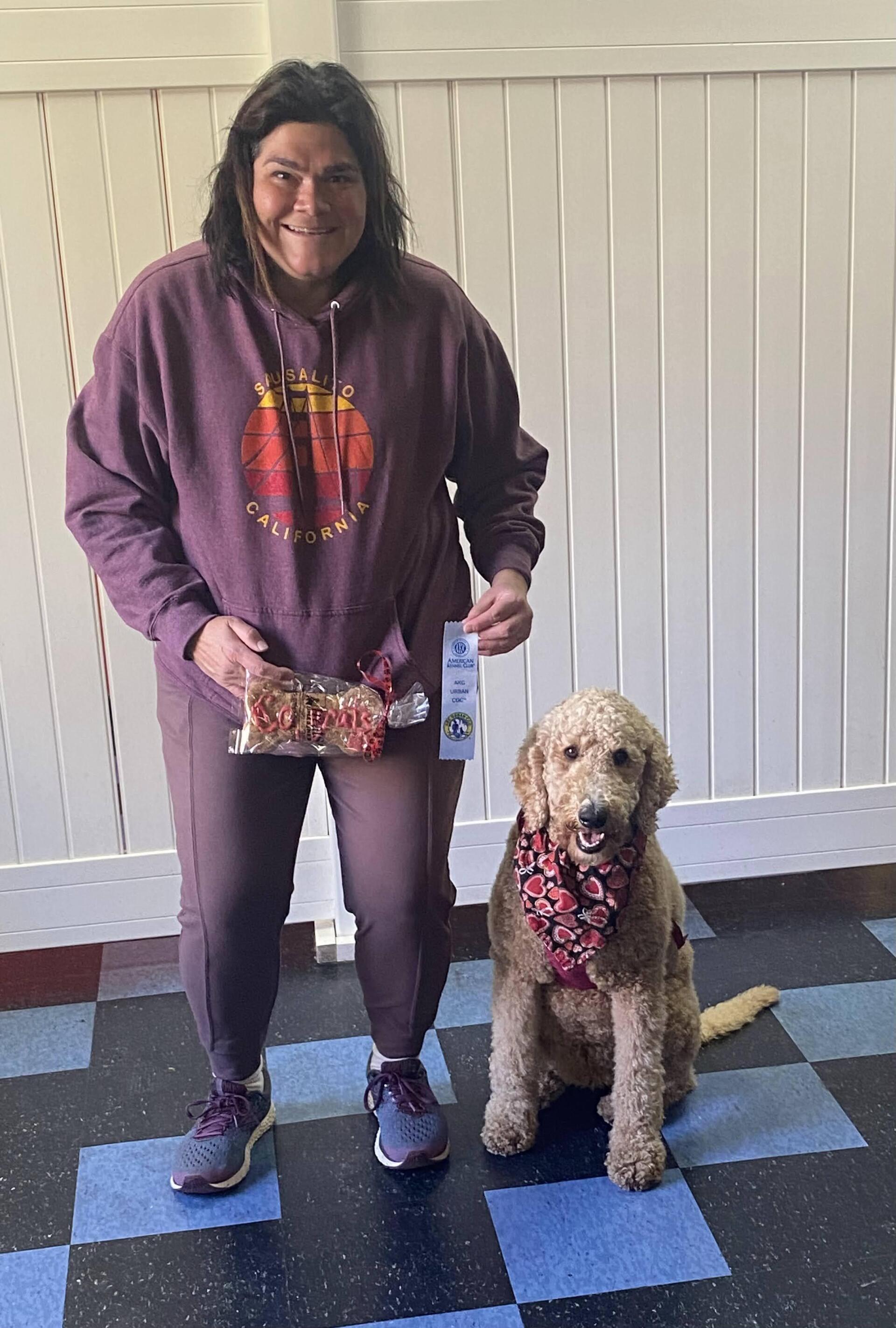 Woman in maroon sweatshirt and pants, holding dog treats, stands with a golden doodle wearing a red bandana; indoors.