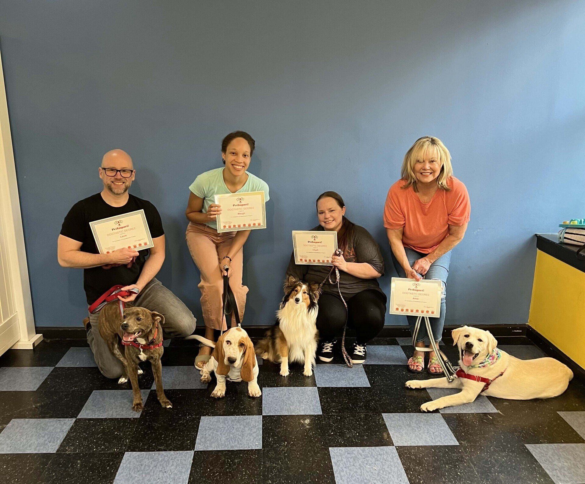 Four people and their dogs pose with certificates indoors. Dogs are various breeds. People smile. Black and blue checkered floor.
