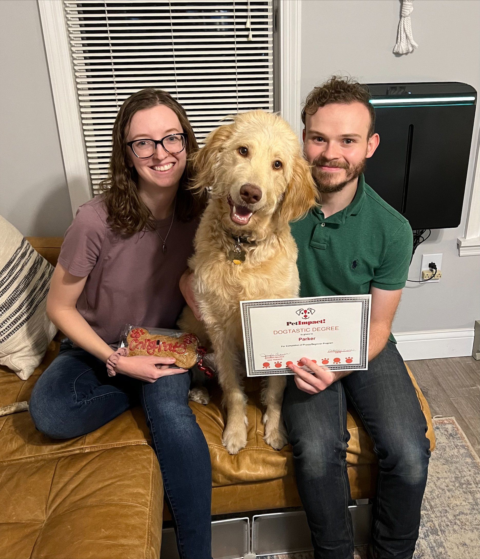 Couple and golden doodle dog on a brown couch, holding a framed certificate.