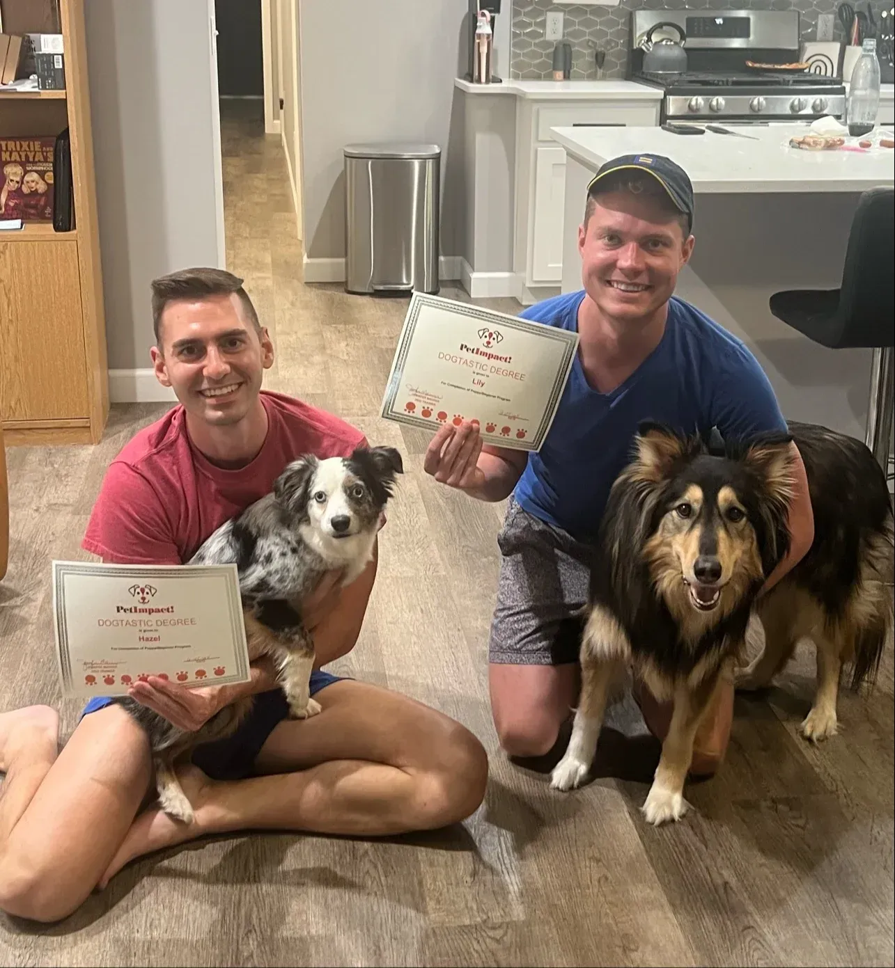 Two men sit with two dogs, holding certificates in a home setting.