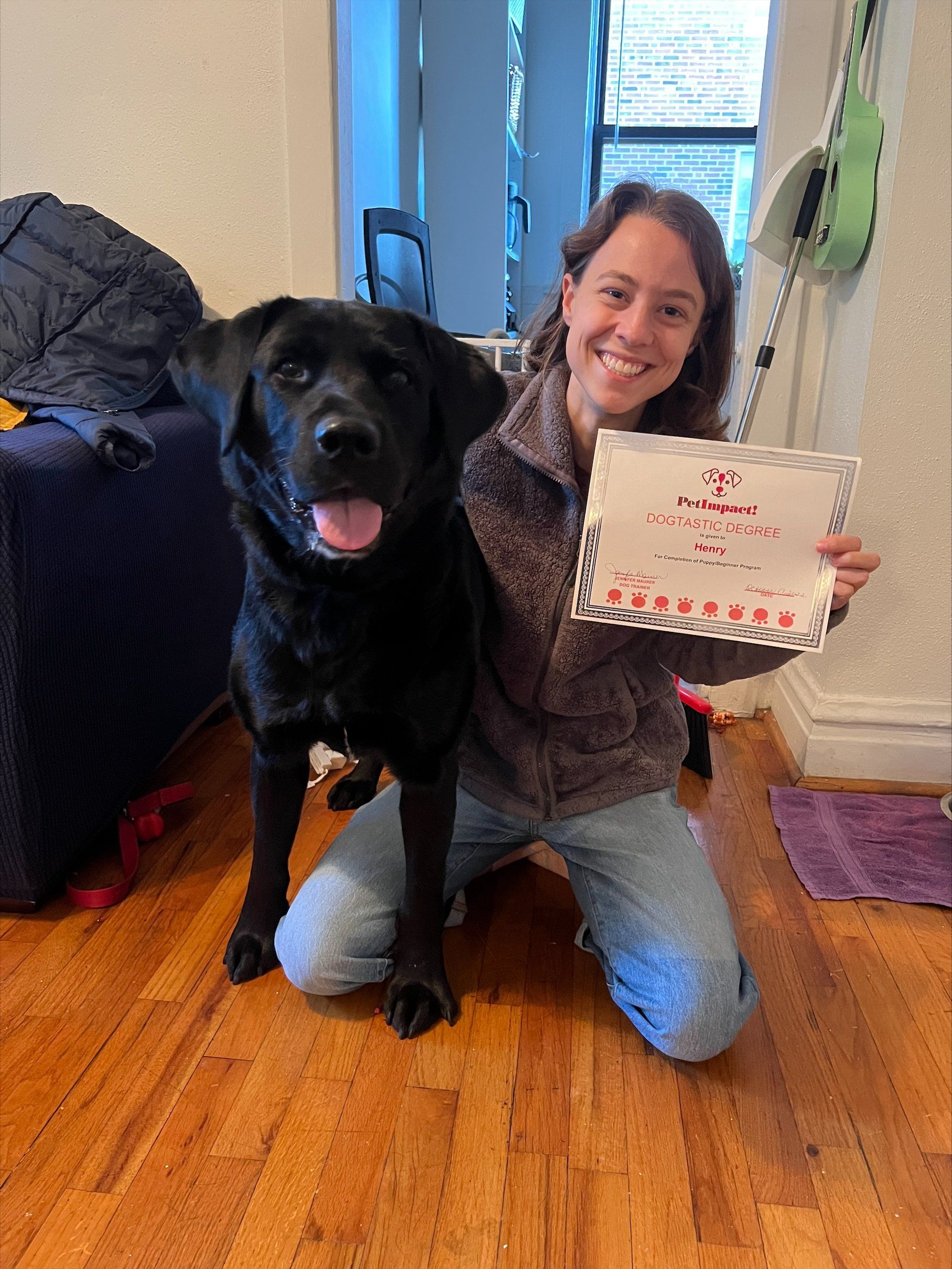 Black dog with its owner kneeling, holding a certificate, smiling. Wooden floor, indoor setting.