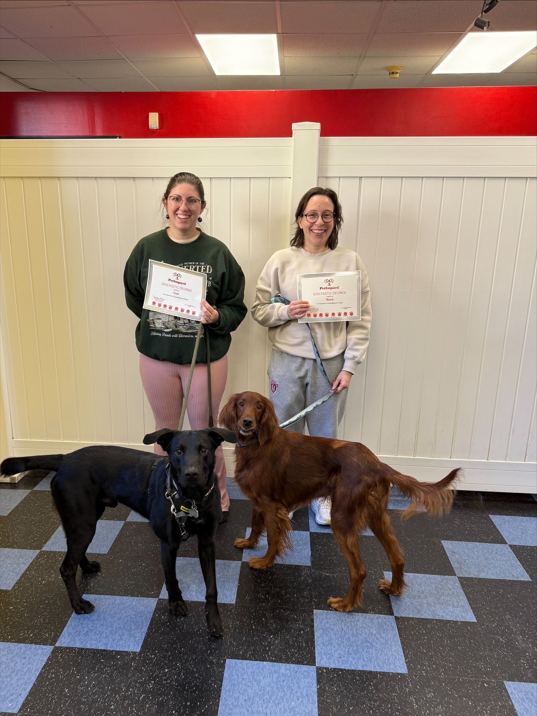Two women hold certificates while two dogs stand in front of them in a tiled indoor room.