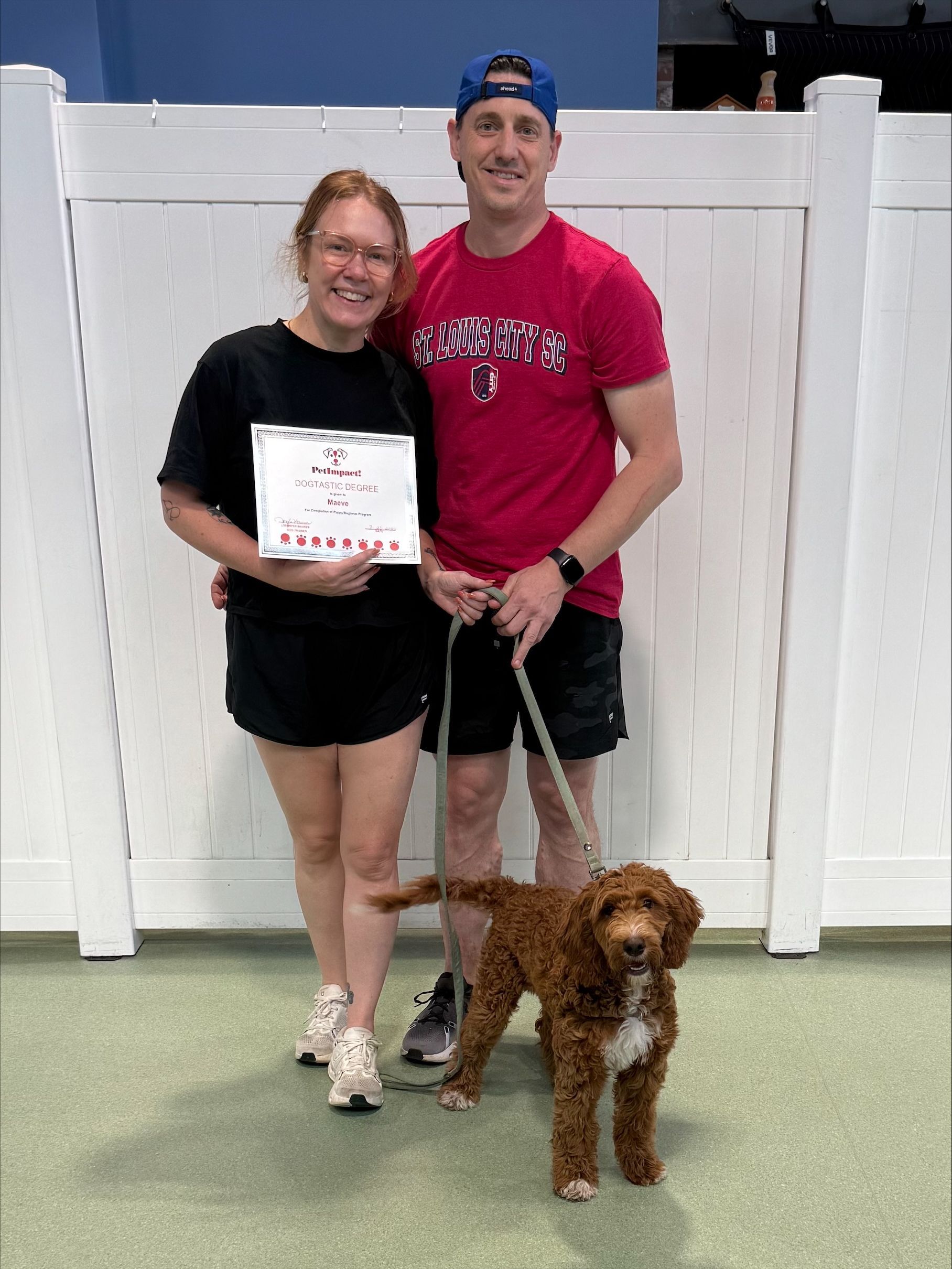Two people with a brown dog standing in front of a white fence, one holding a certificate.