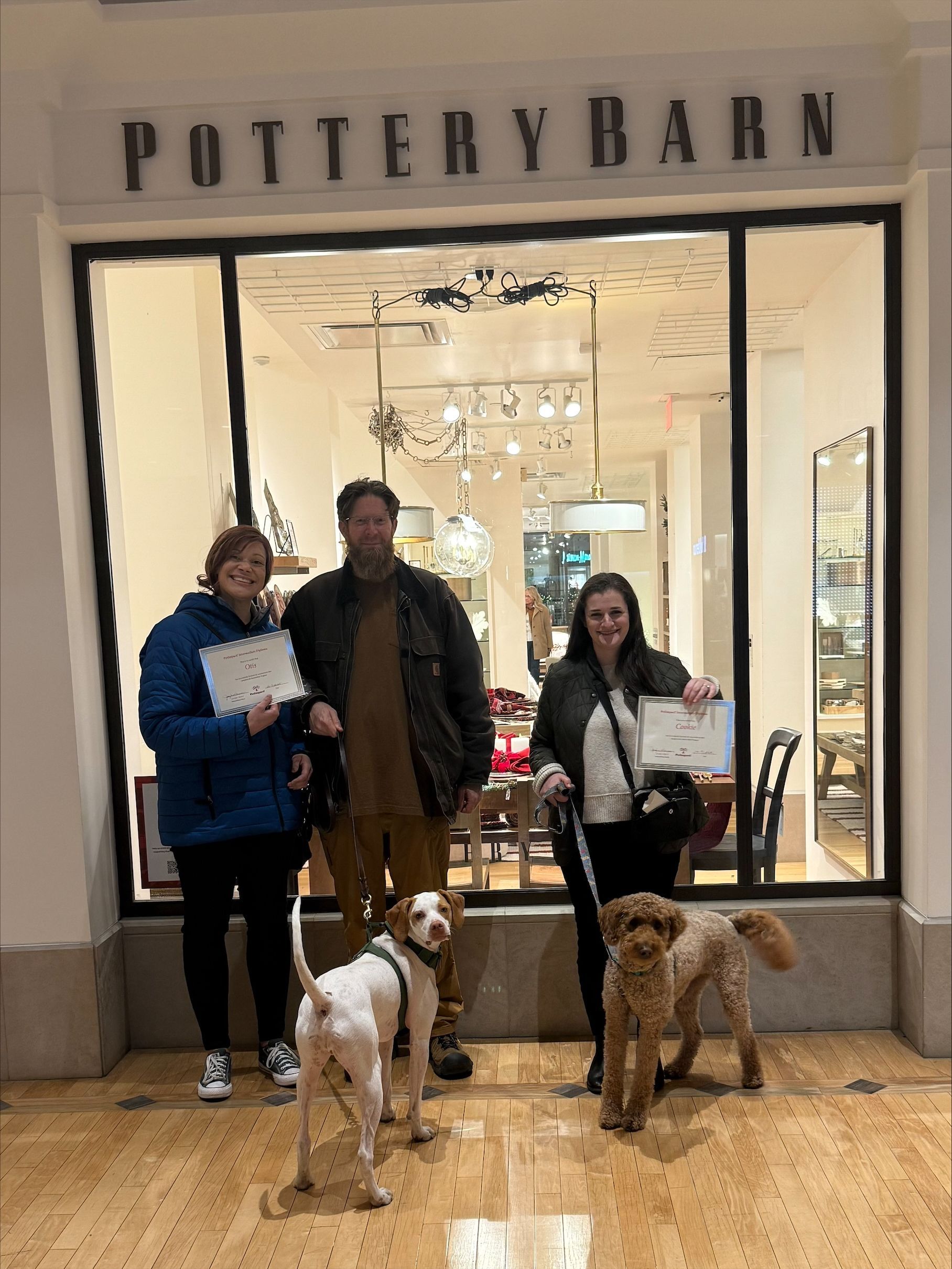Three people with two dogs pose outside a Pottery Barn storefront.