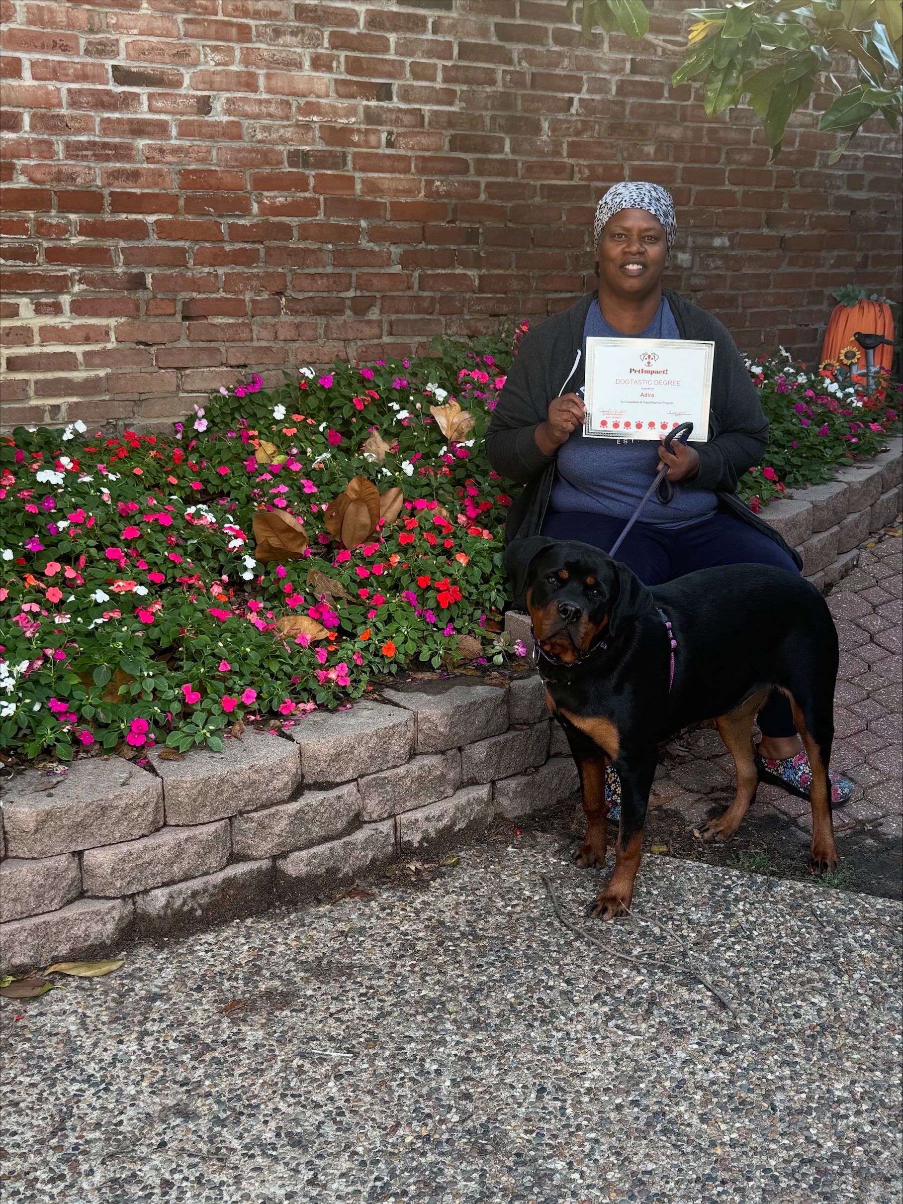 Woman with dog, holding a certificate. Brick wall backdrop, flower bed in front.