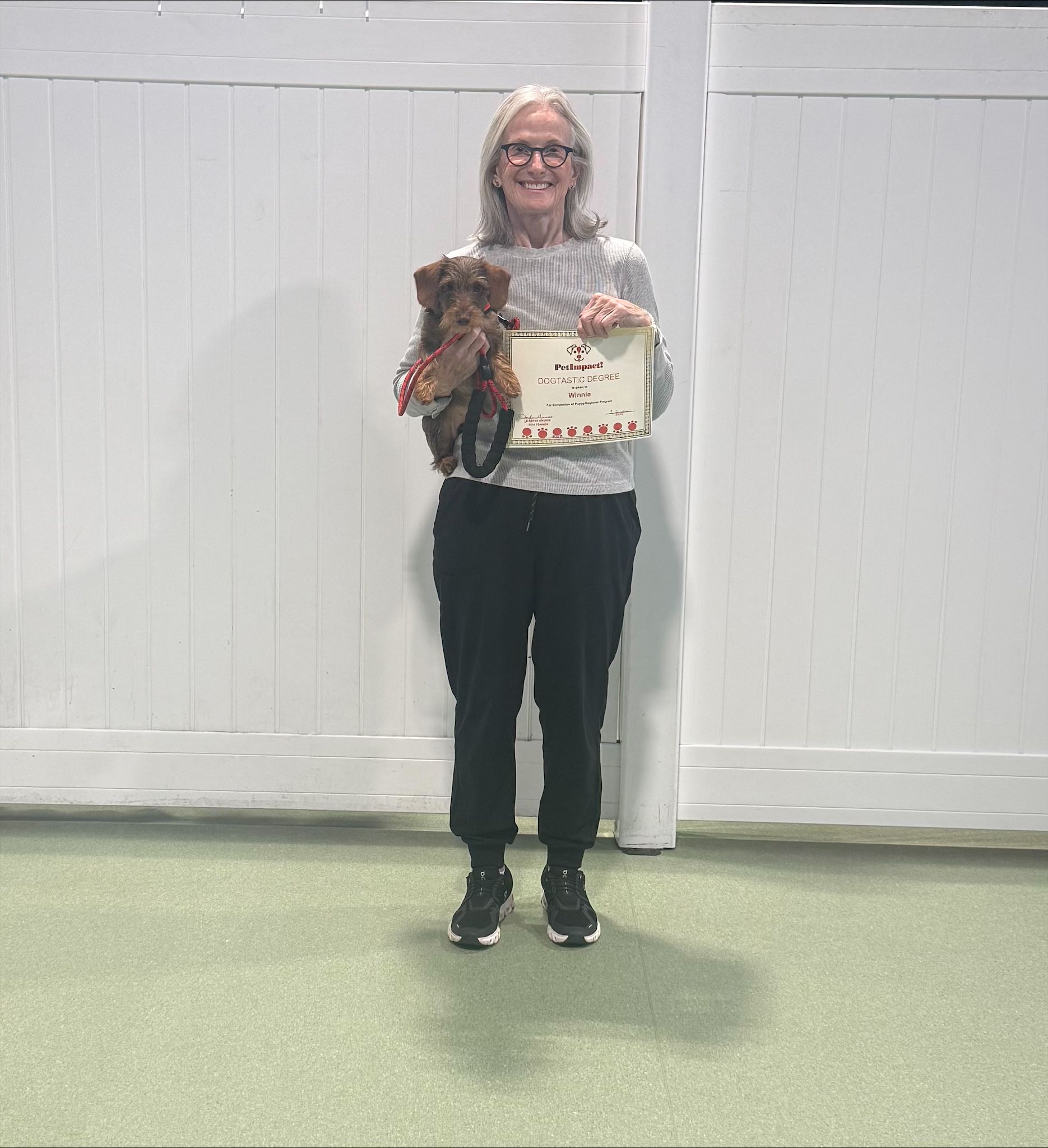 Person holding a small dog and a framed certificate, standing indoors against a white wall.