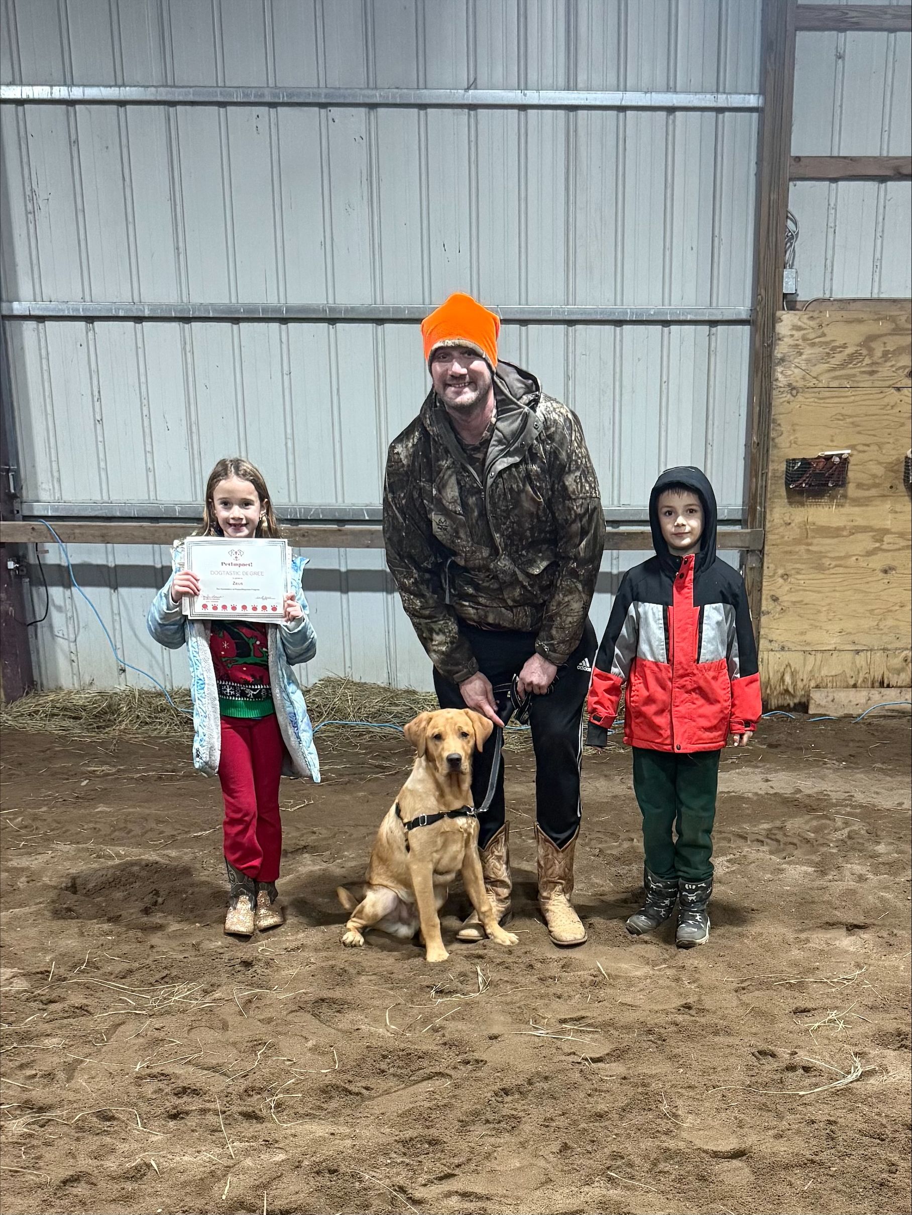 Three people stand with a dog in a muddy barn, holding a sign and red bags.