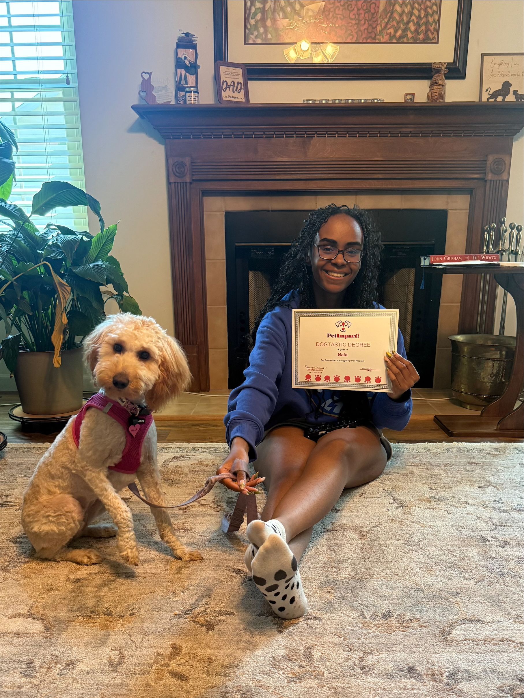 Person sitting on carpet with a small dog, holding a framed certificate in front of a fireplace.