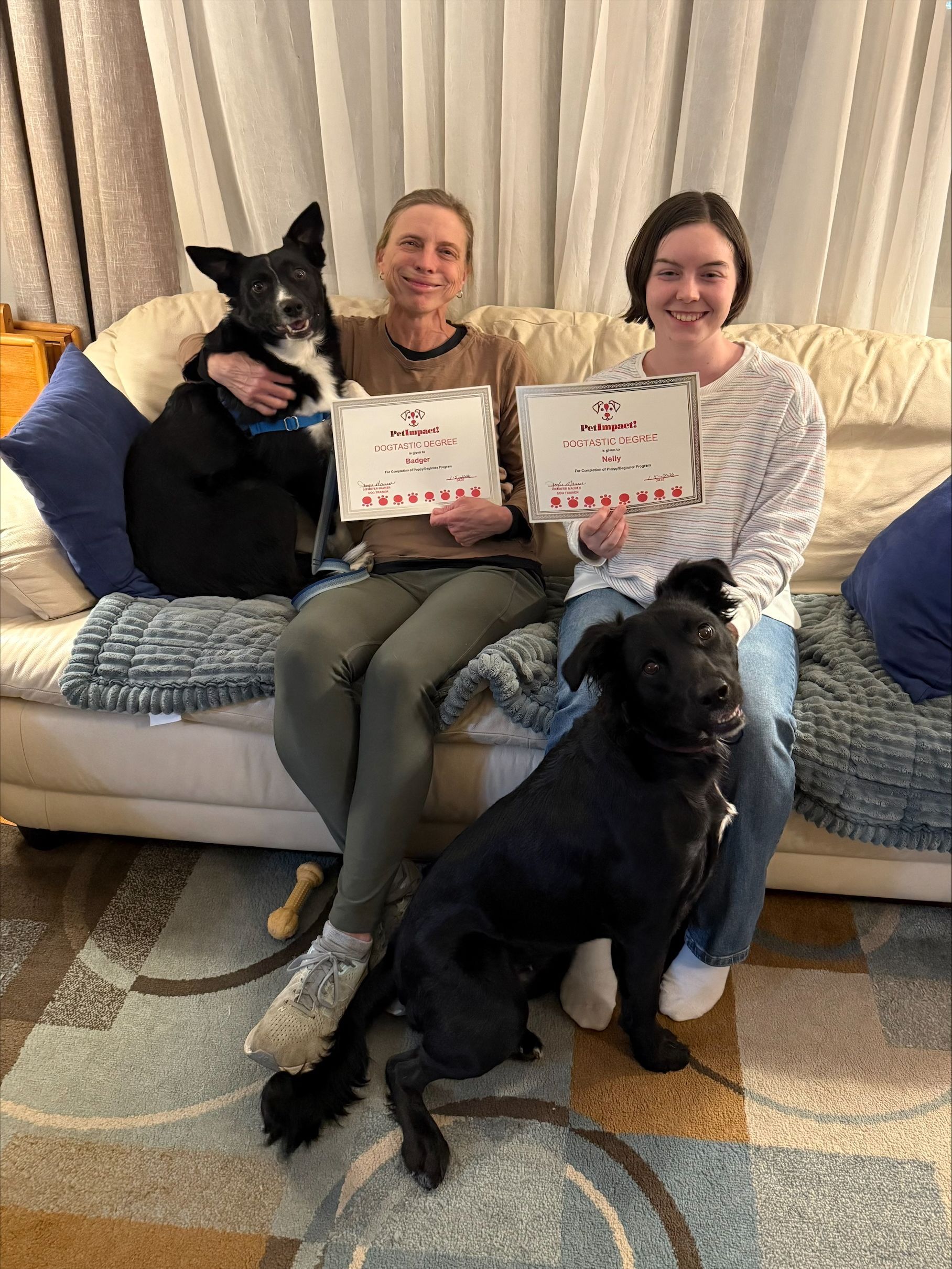 Three people with two dogs on a couch, holding award certificates in a cozy living room.