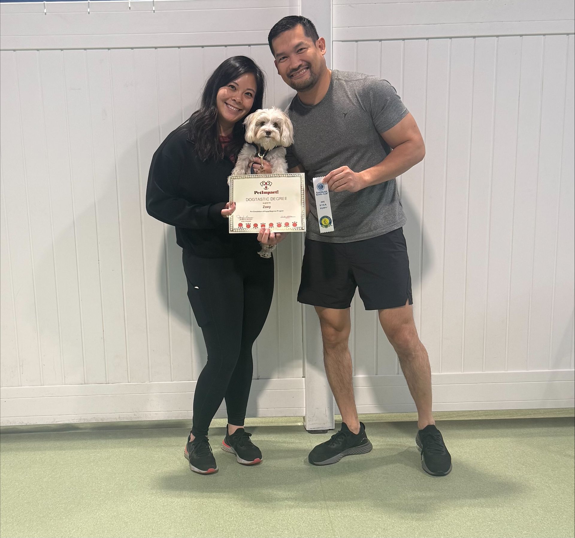 Two people posing with a small dog and a certificate against a white wall