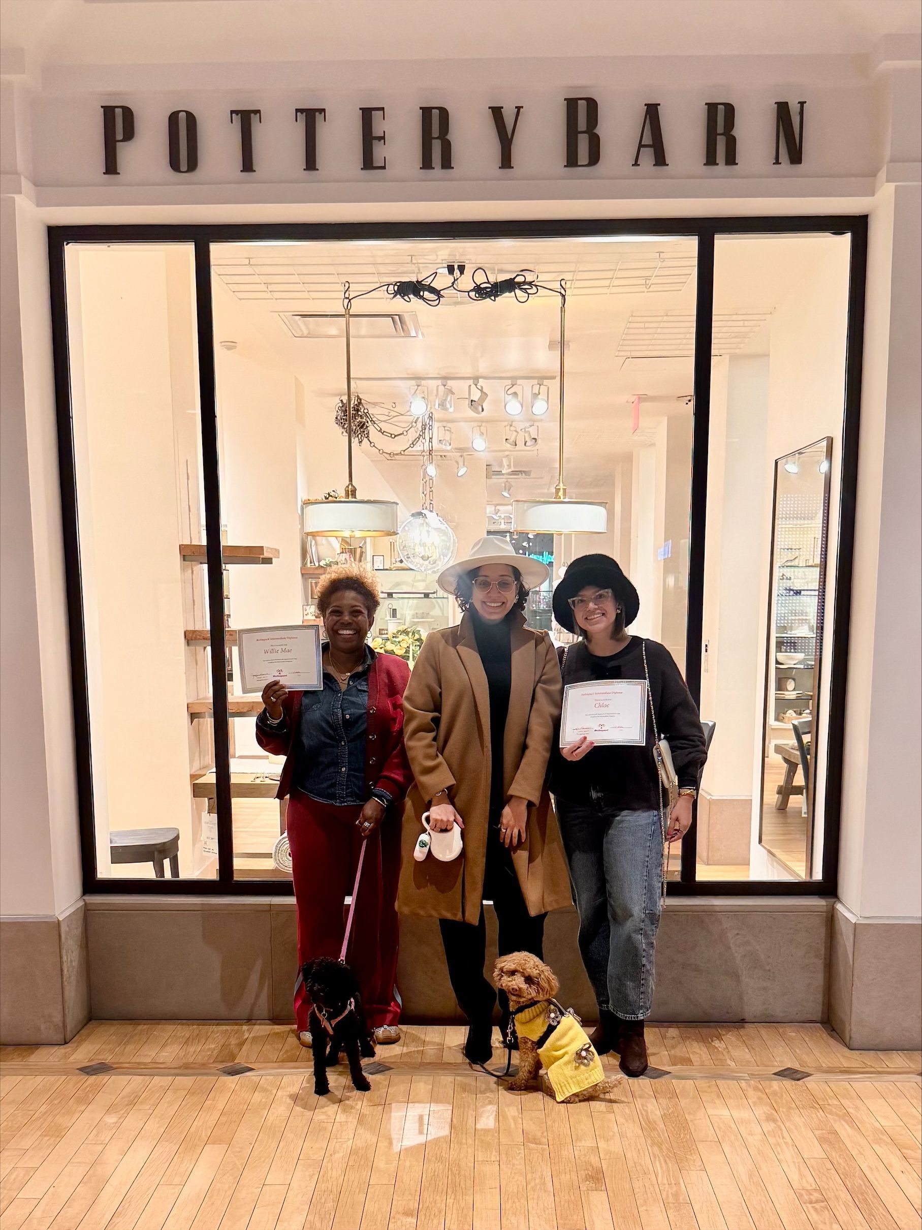 Three people pose outside the Pottery Barn store, with shopping bags and a small dog at their feet.