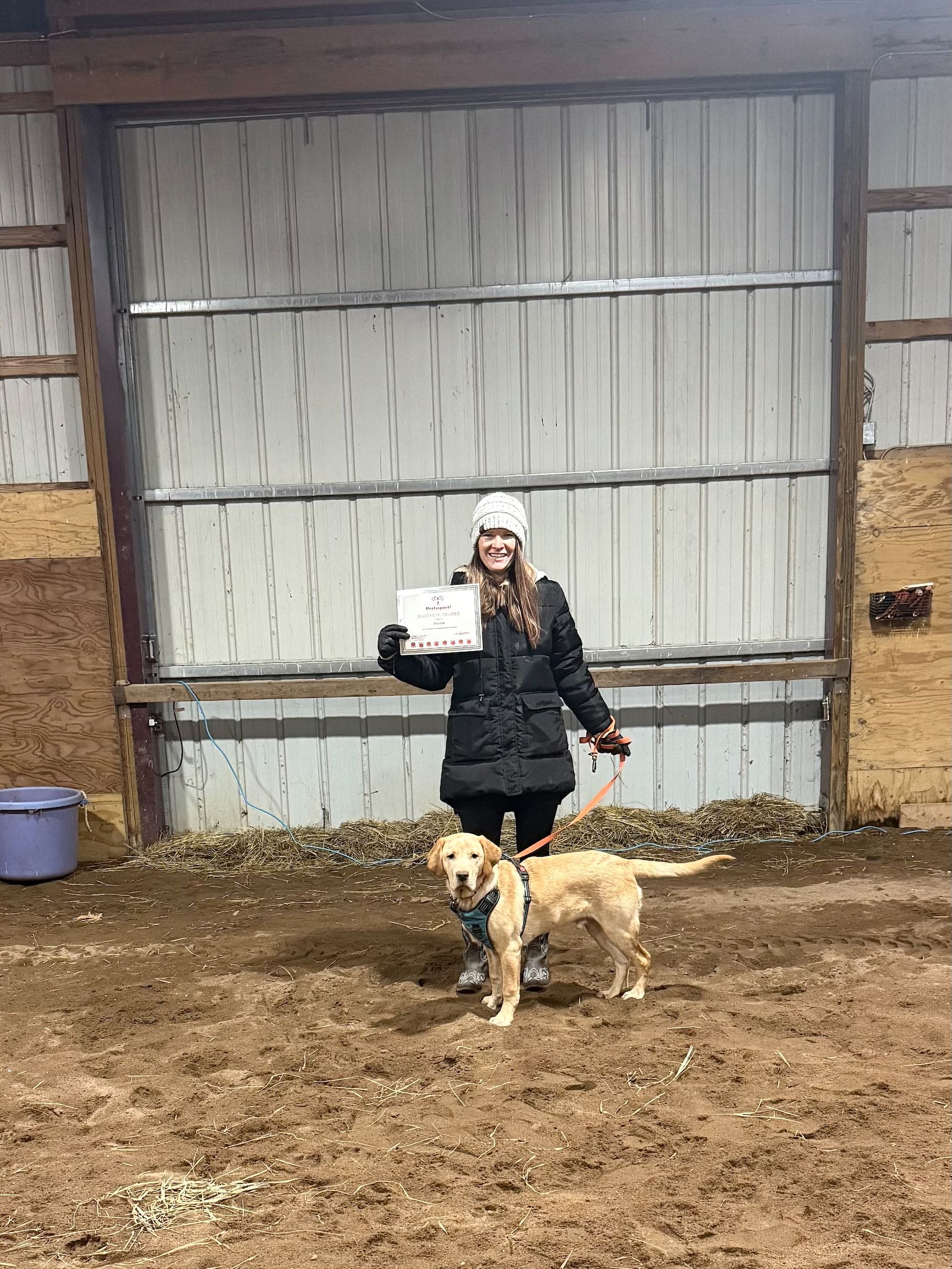 Person standing with a tan dog in an indoor barn-like space, holding a small white animal.