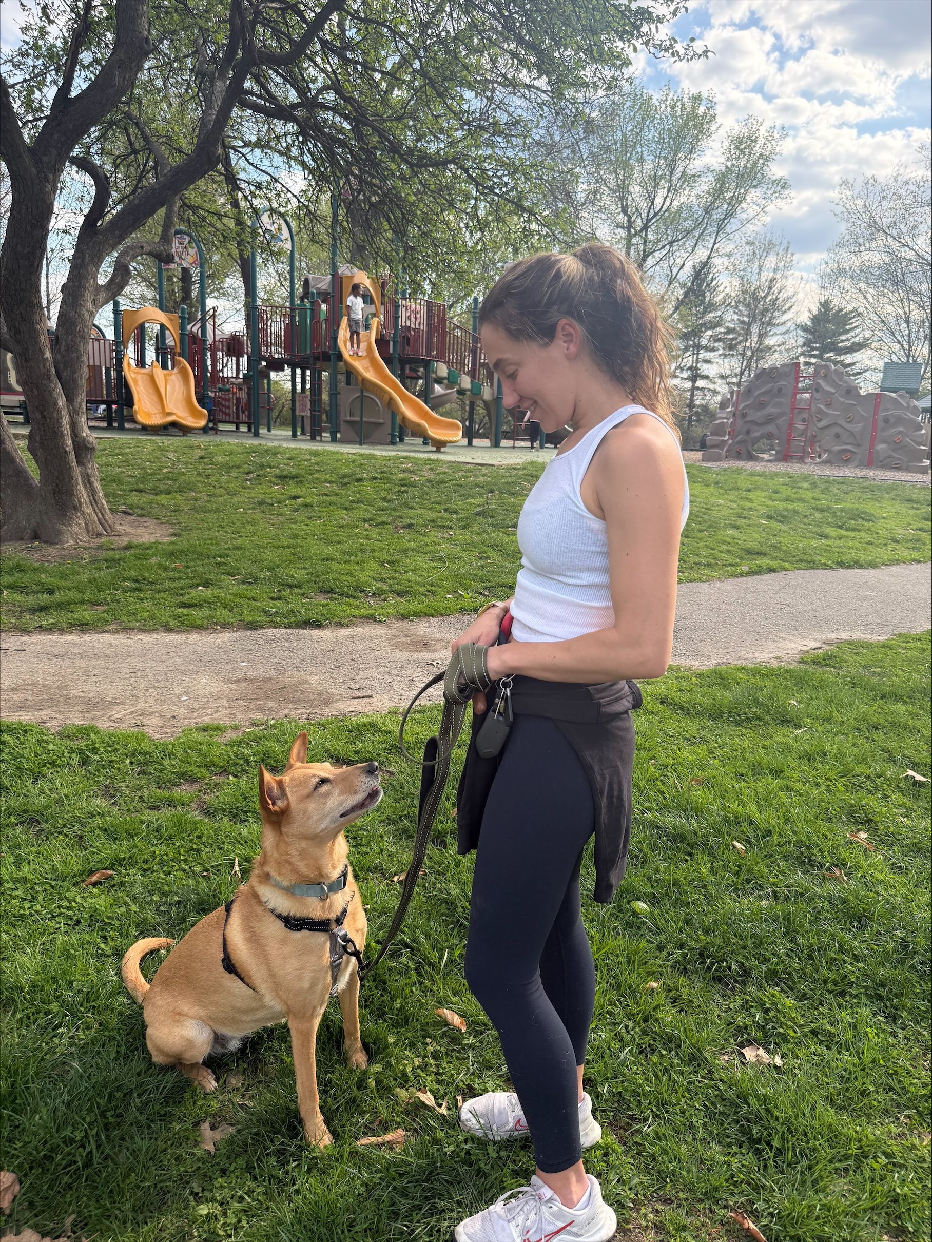 Woman in white tank top and black leggings with a tan dog on a leash in a park near playground slides.