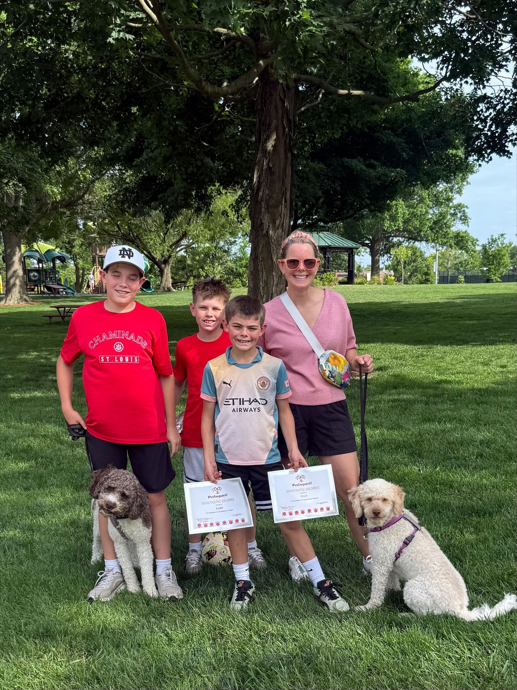 Four people and two dogs standing on grass under a tree in a park, holding signs and flowers