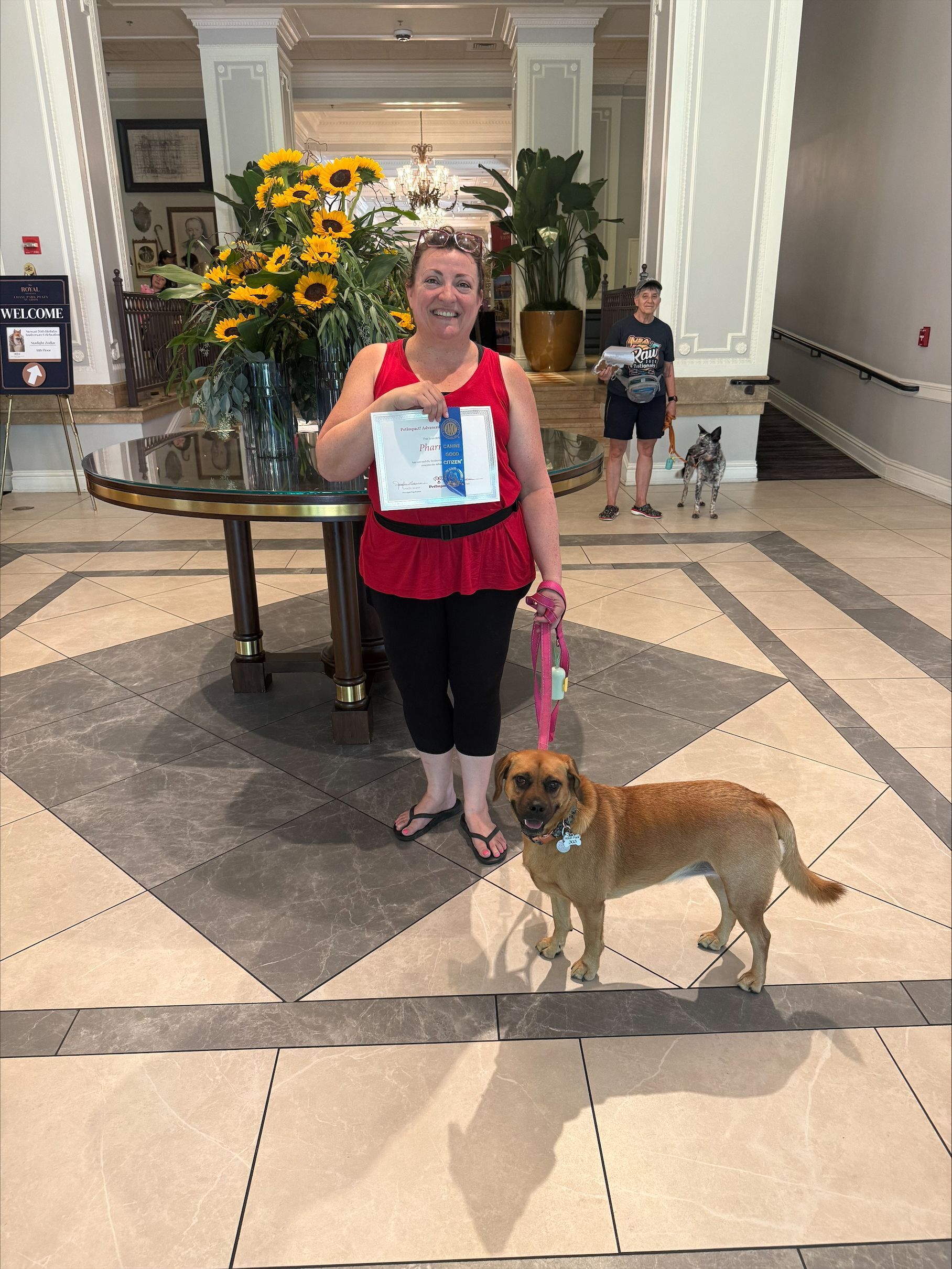 Woman holding a sign and leash with a dog in a hotel lobby beside a large sunflower arrangement
