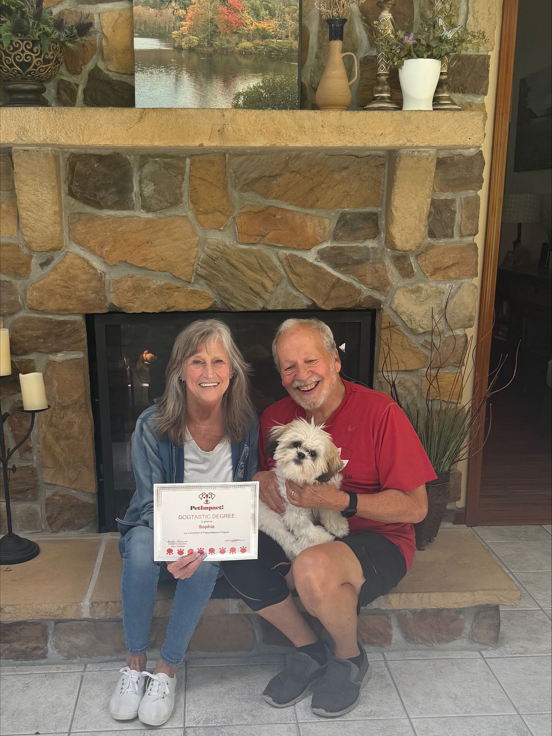 Two people seated by a stone fireplace, holding a small dog and a certificate, smiling at the camera