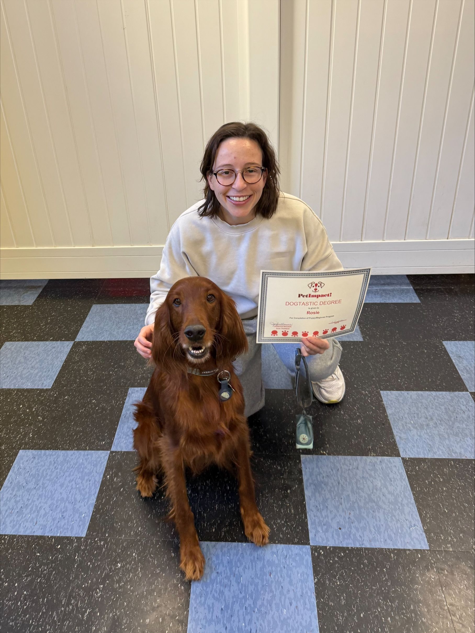 Person kneels with a brown dog and a certificate on a checkered floor indoors
