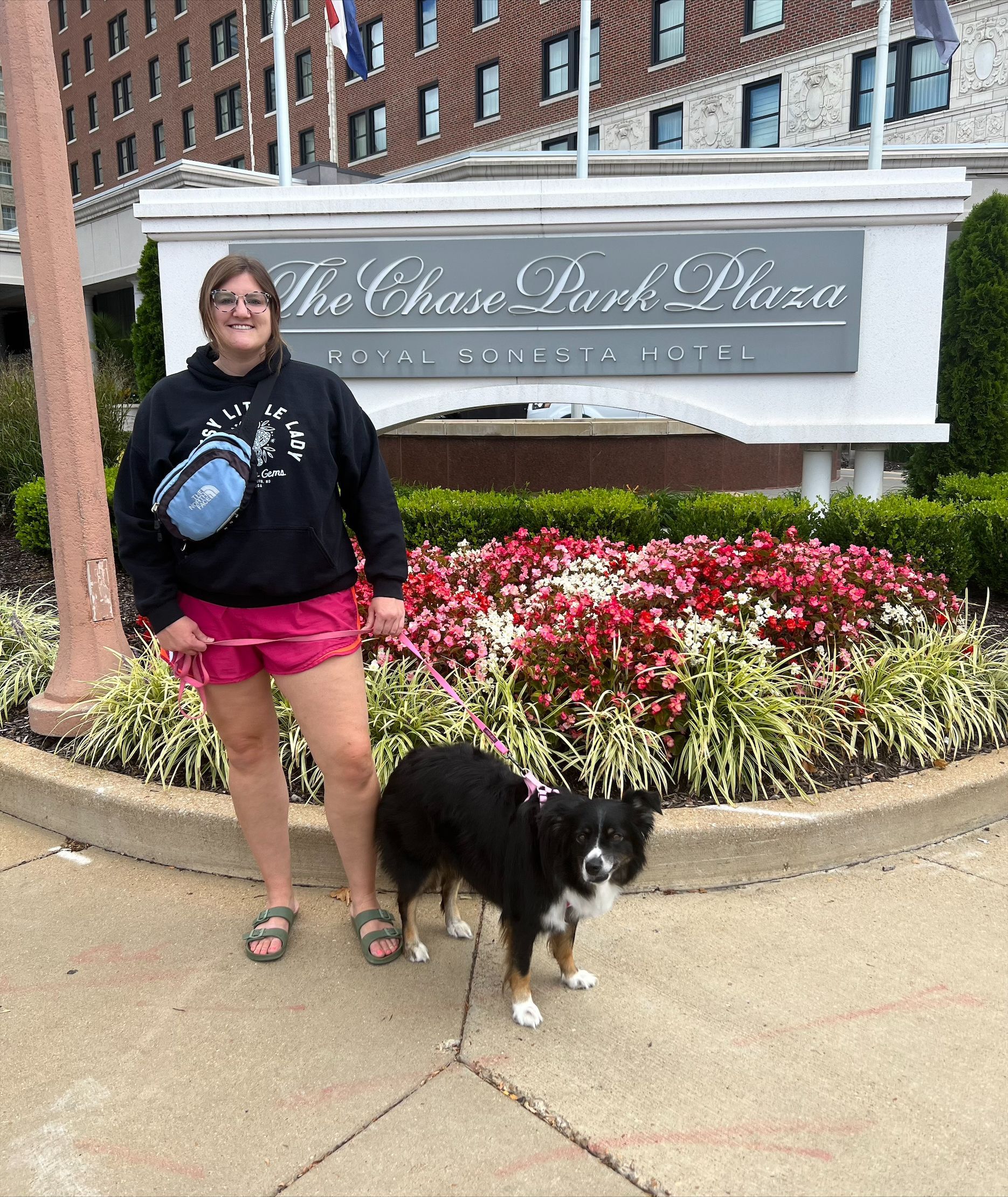 Woman and dog pose in front of the Chase Park Plaza hotel sign. Woman wears shorts and sweatshirt; dog is black and white.
