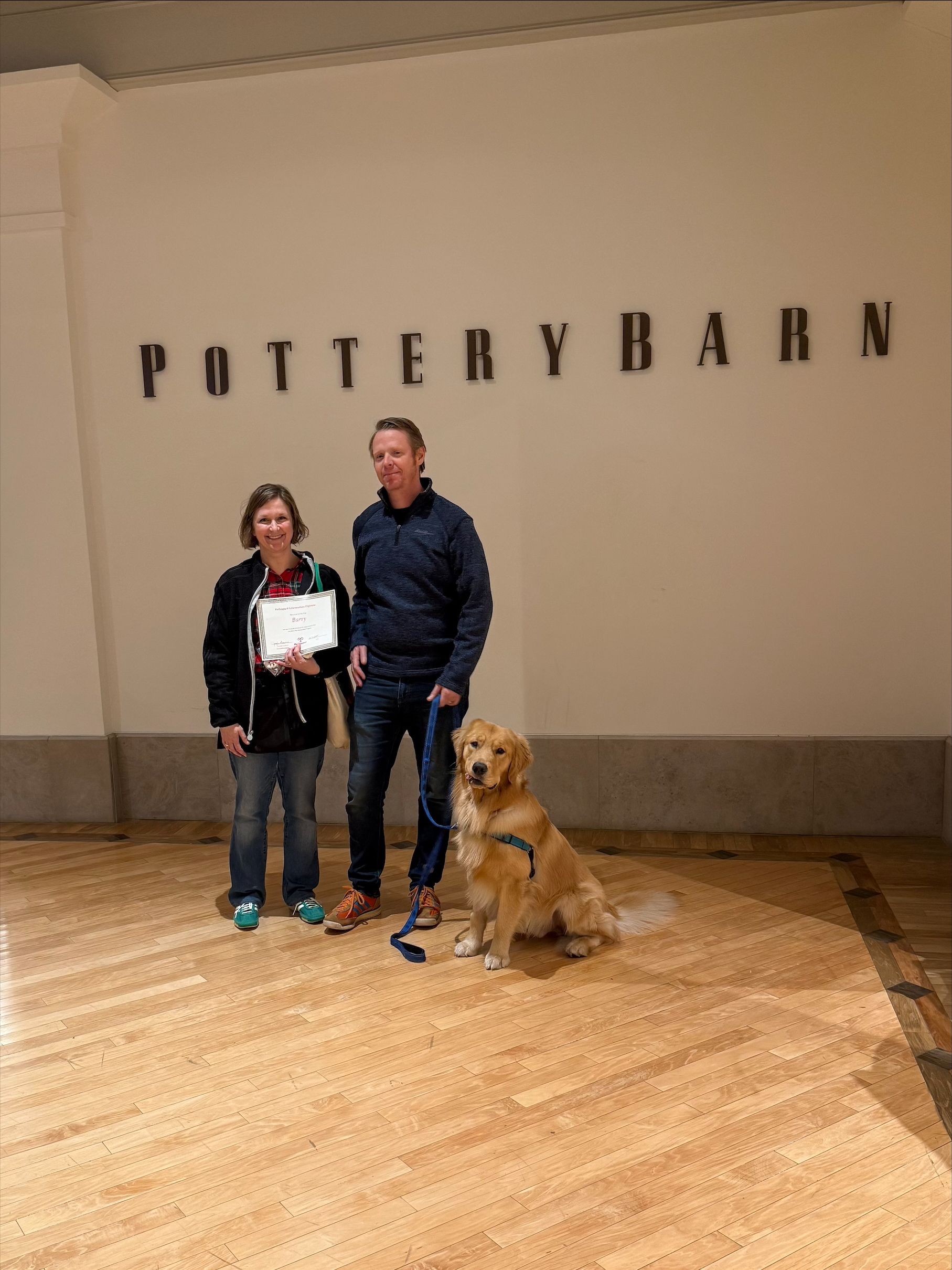Two people posing with a golden retriever in front of a Pottery Barn sign.