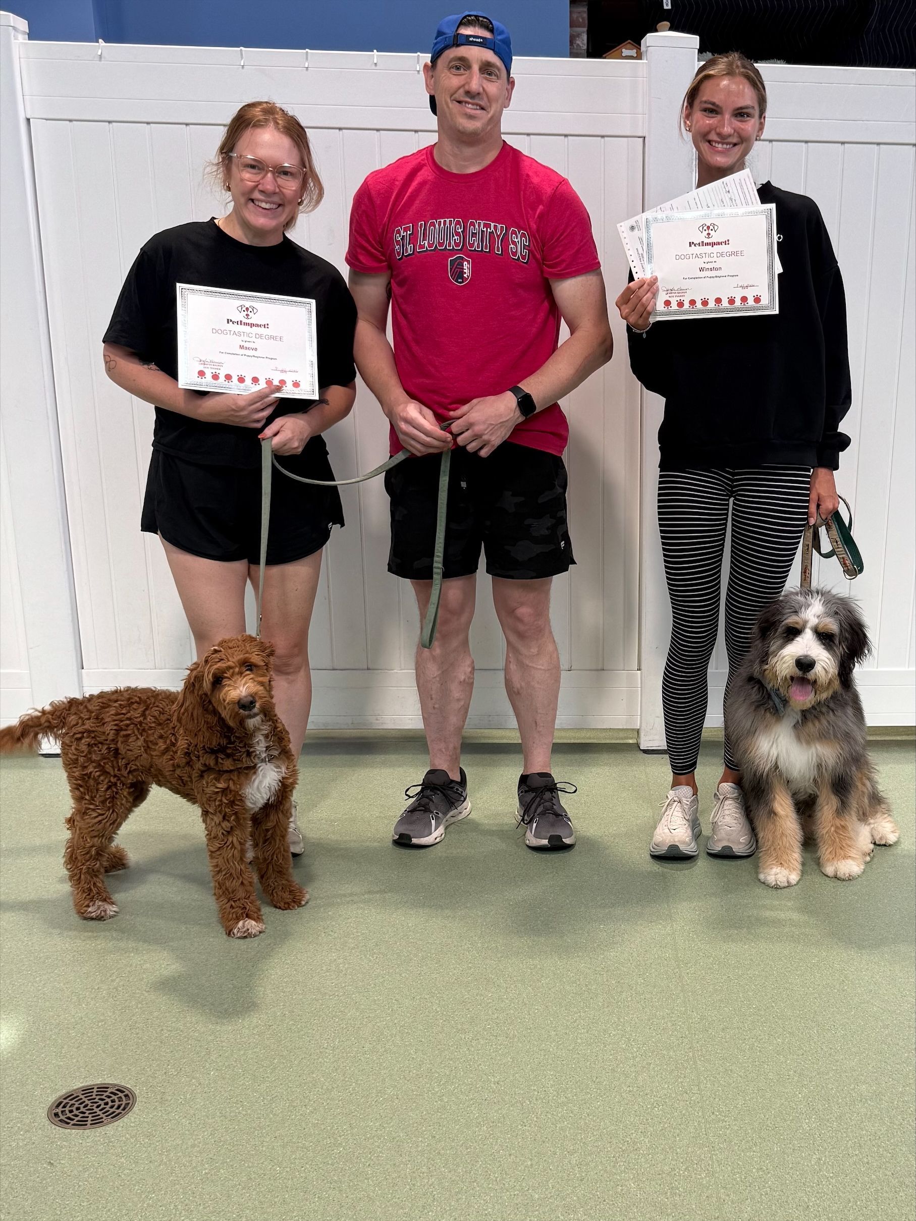 Three people hold certificates and leashes with two dogs in an indoor training room.