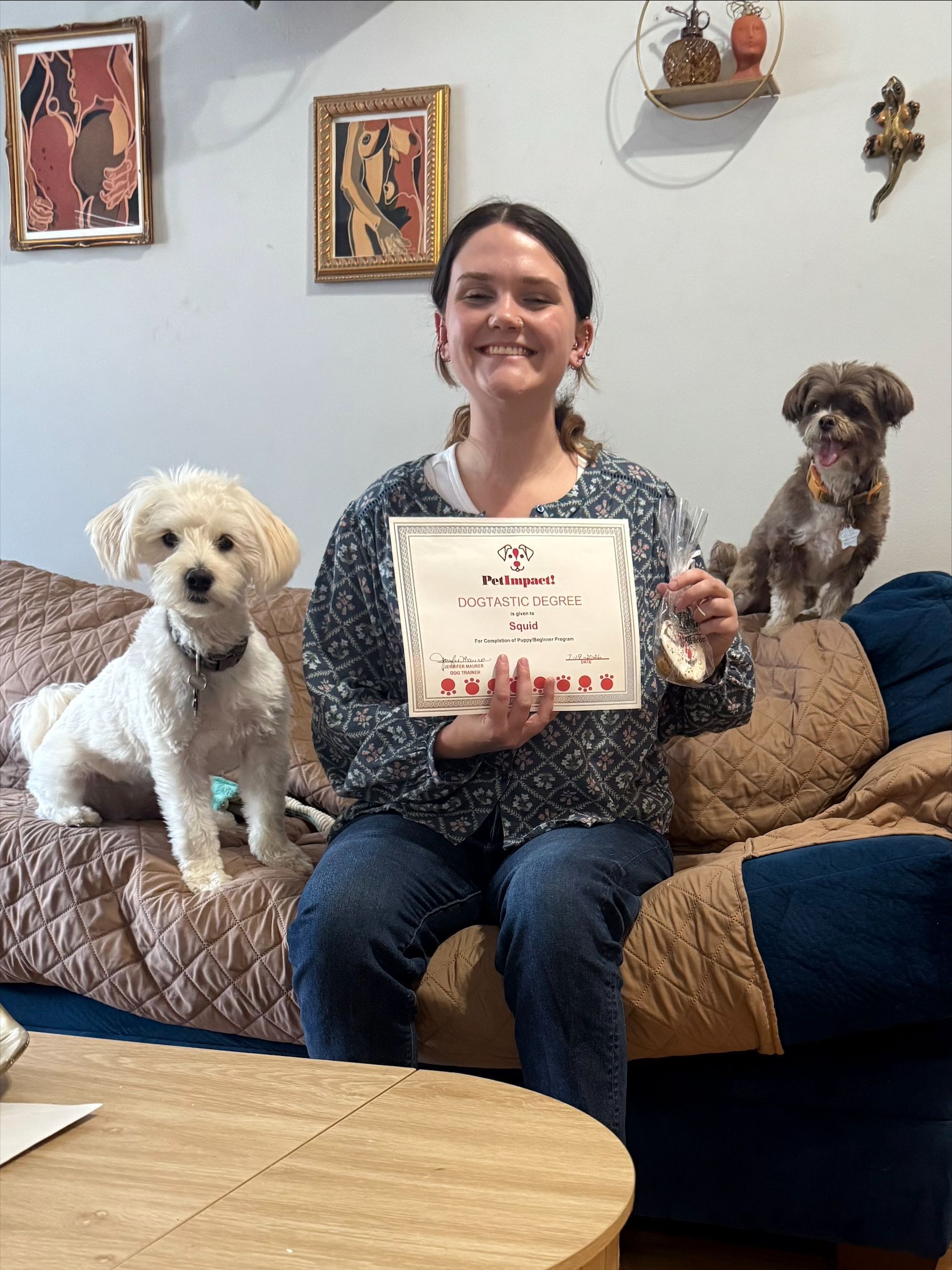 Woman holding a certificate, seated on a couch with two small dogs in a cozy living room
