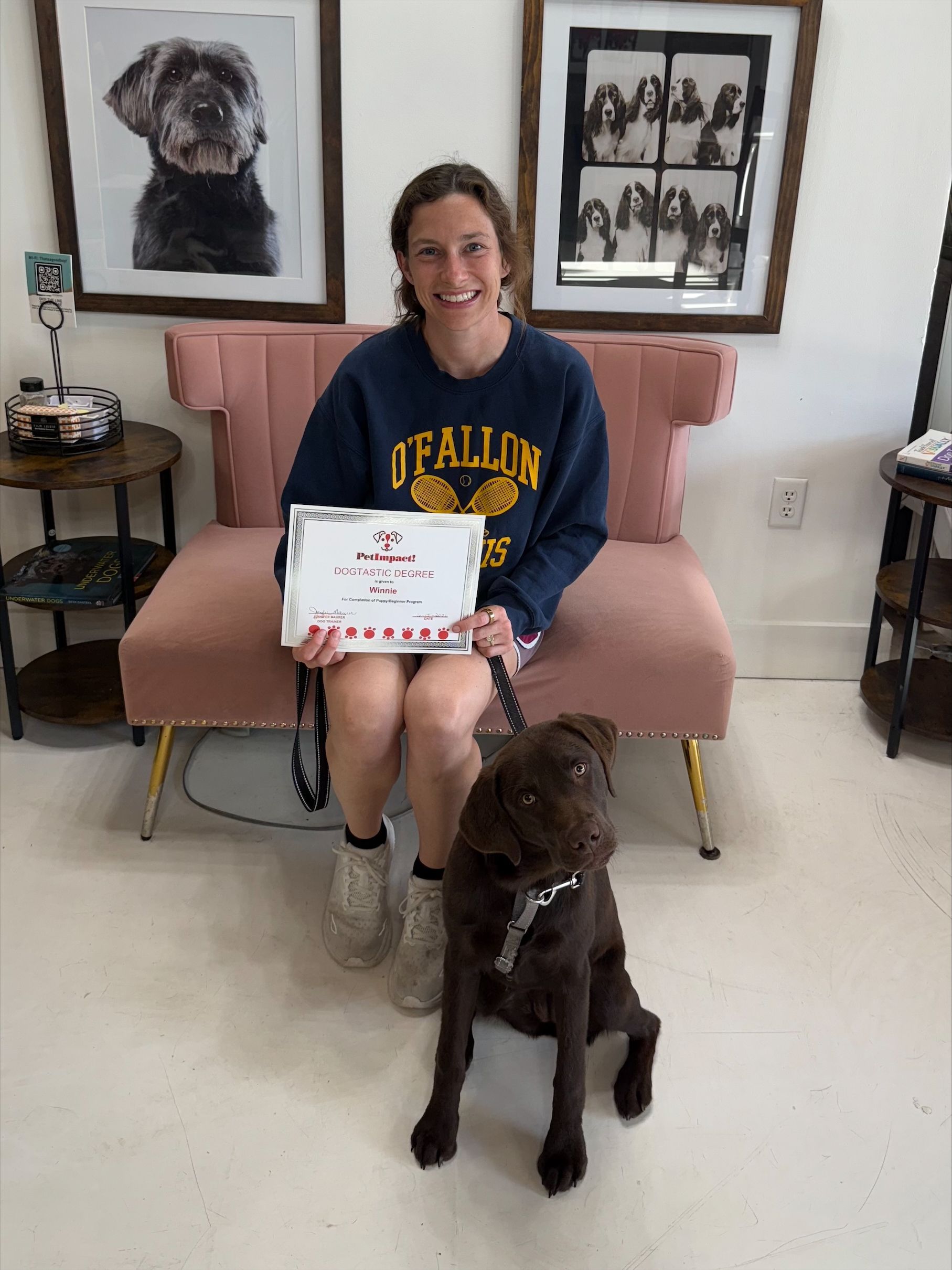 Woman holding a certificate sits on a pink couch with a brown dog in front of her, framed dog portraits behind.