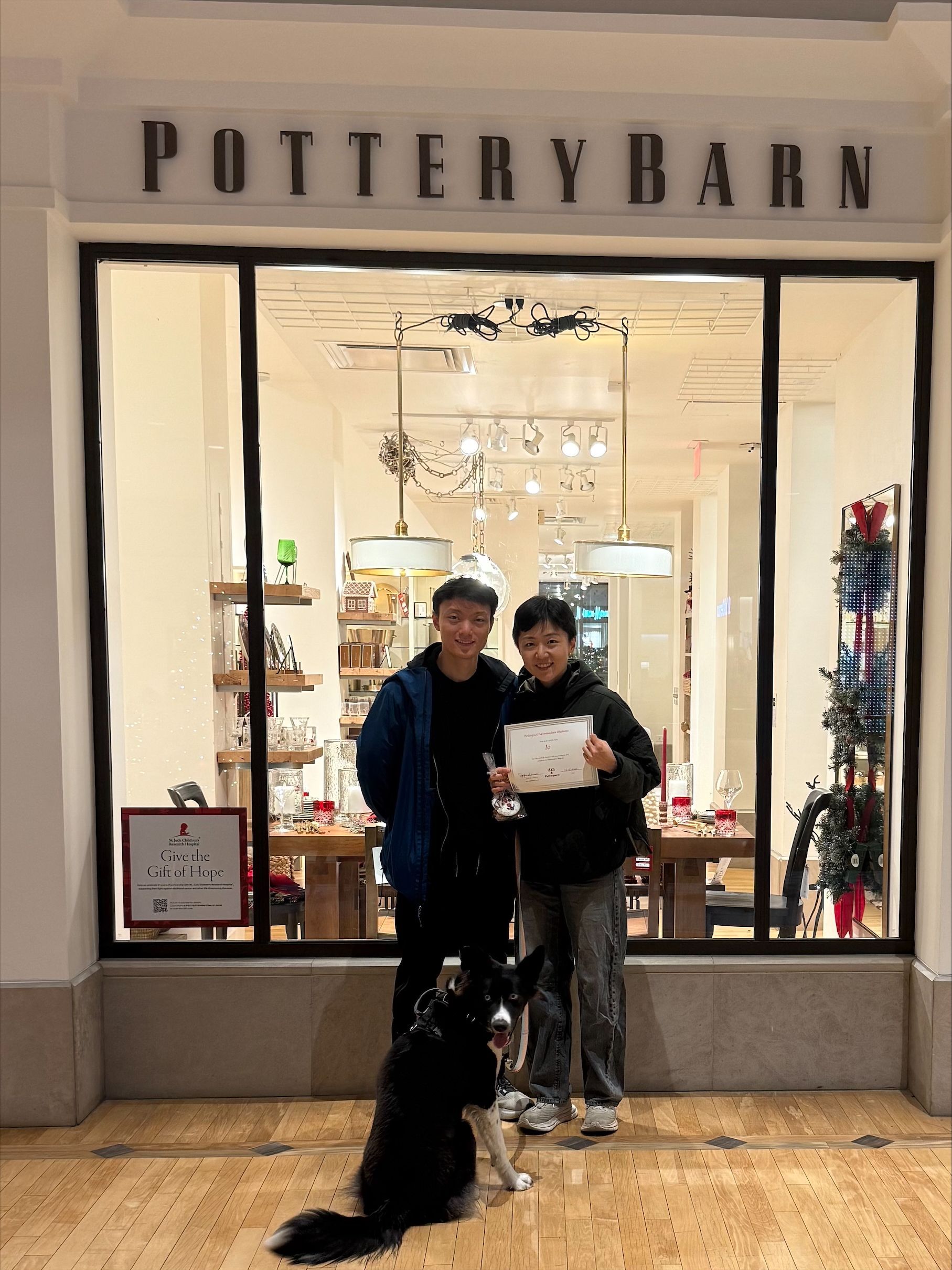 Two people and a black dog stand at the entrance of a Pottery Barn store decorated for the holidays.