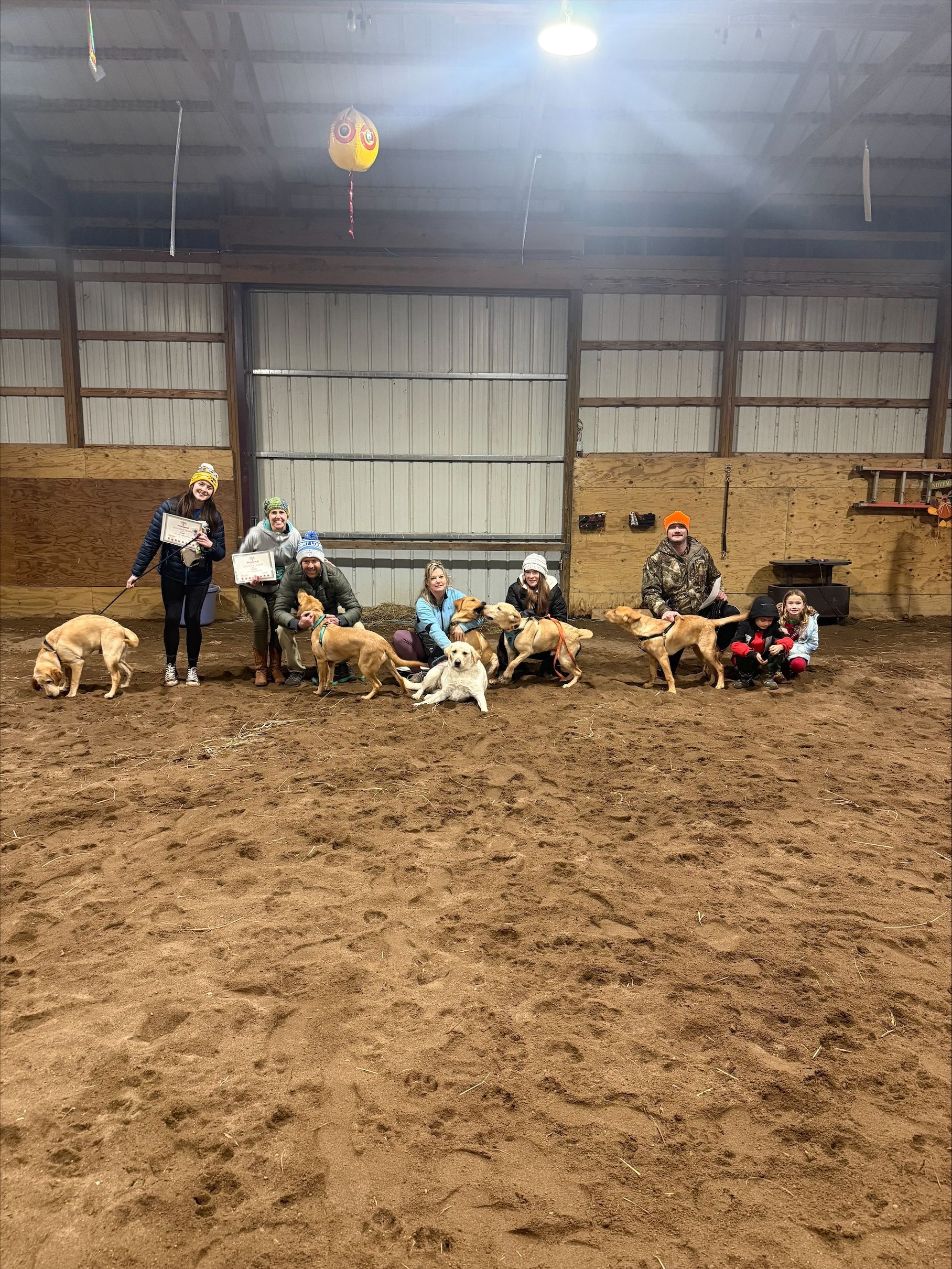 Children and goats gathered in an indoor barn, with people standing and sitting on a dirt floor.