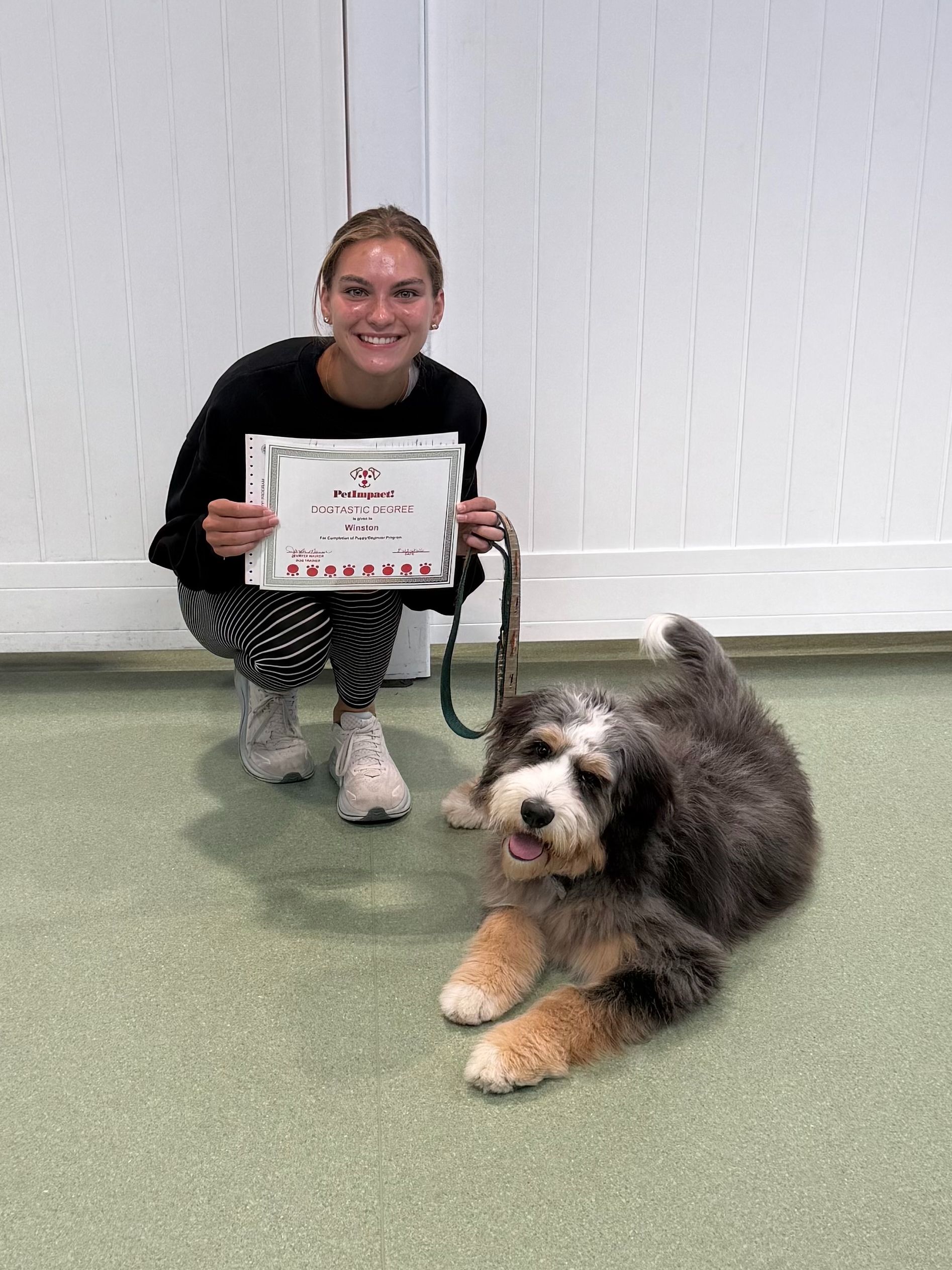 Woman kneeling beside a fluffy dog, holding a certificate in a bright indoor room