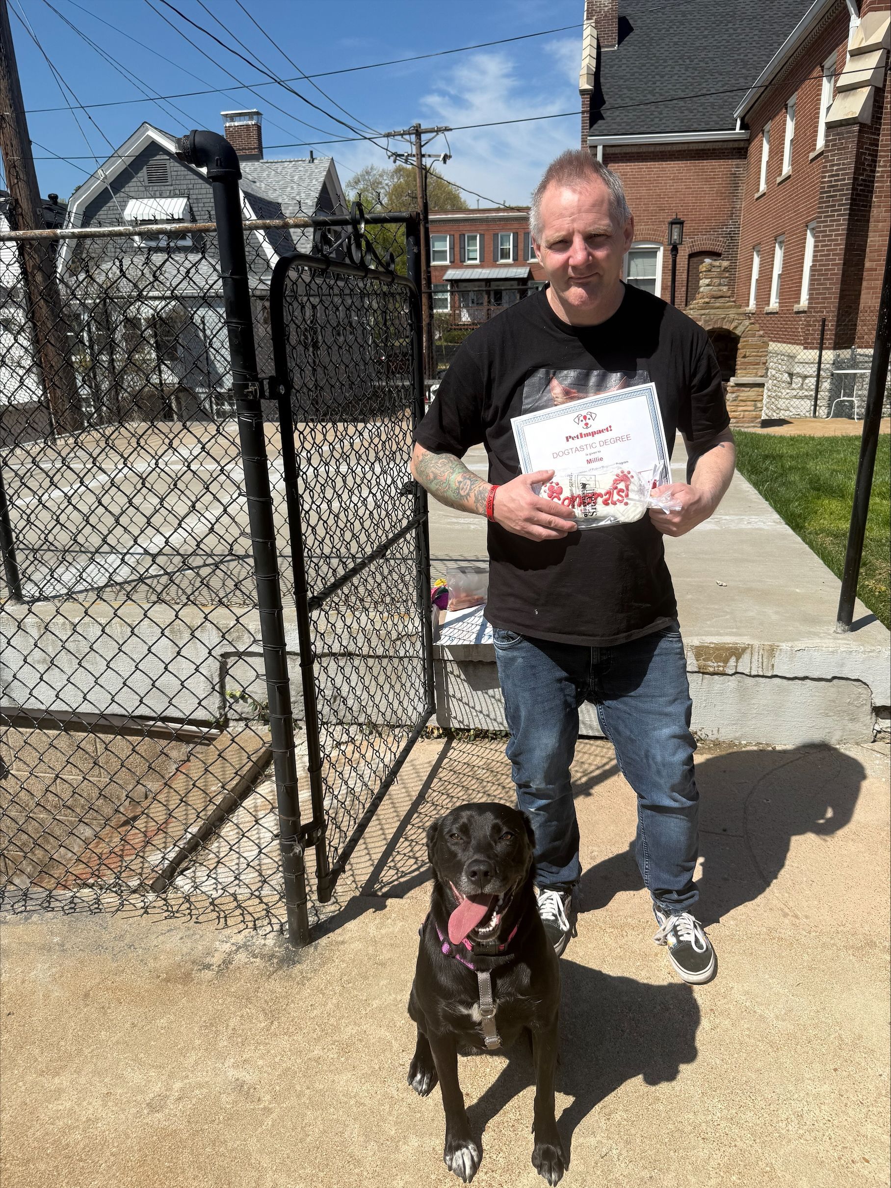 A man stands outdoors on a sunny day holding a certificate next to a black dog with its tongue out.