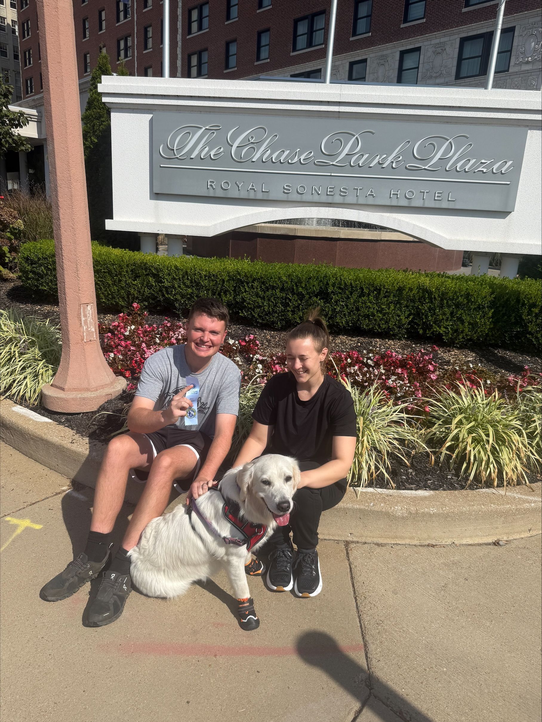 Two people and a white dog pose in front of the Chase Park Plaza Hotel sign.
