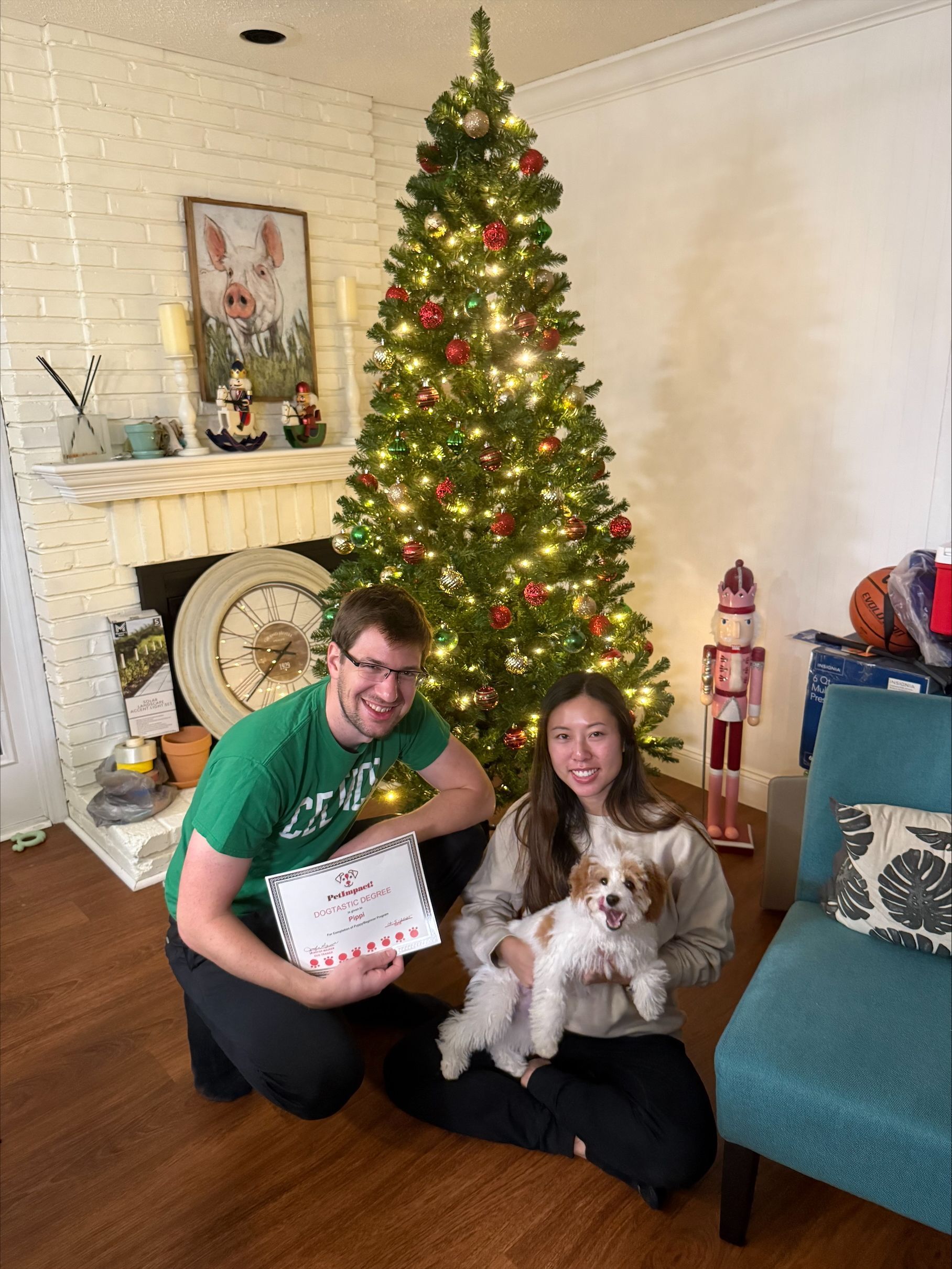 Two people and a small dog pose beside a decorated Christmas tree in a cozy living room.