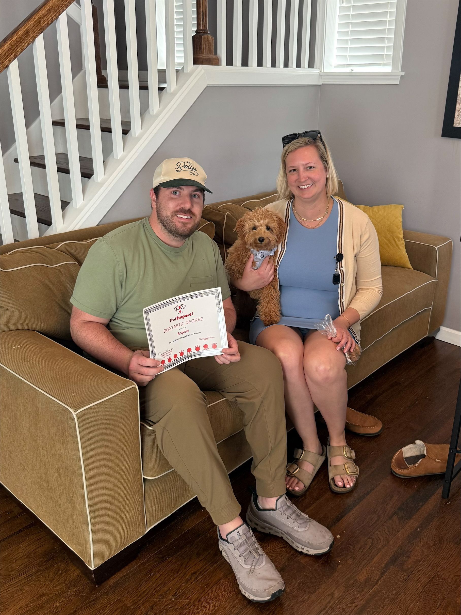 Two adults on a brown couch with a dog, holding a certificate in a bright living room