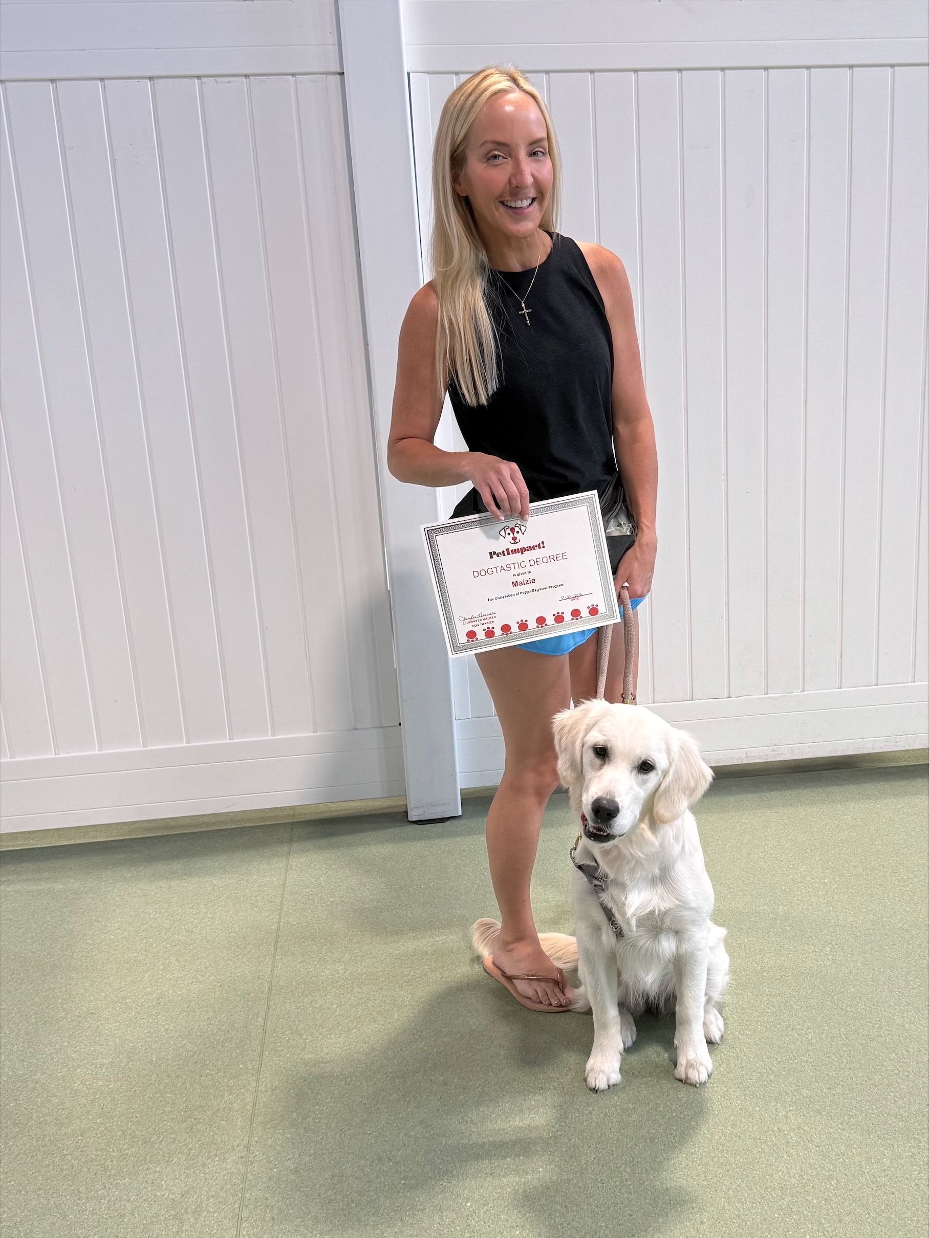 Woman standing indoors with a white dog, holding a framed certificate against a white fence backdrop