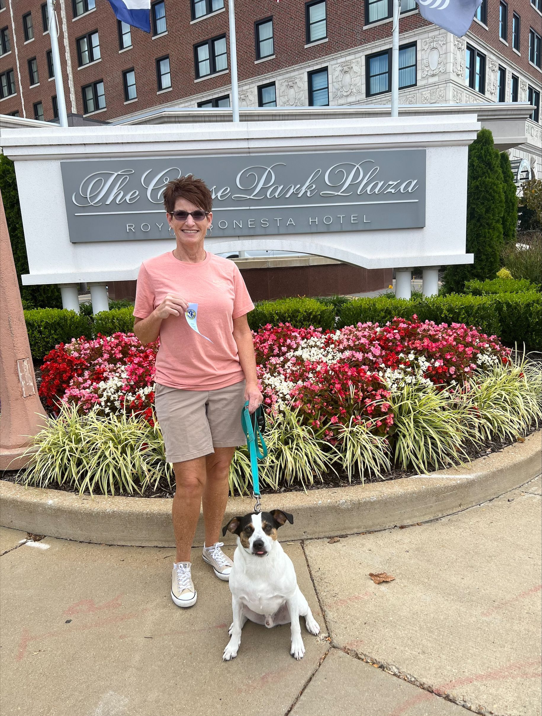 Woman and dog pose in front of a hotel sign. The woman holds a leash, next to a flower bed.