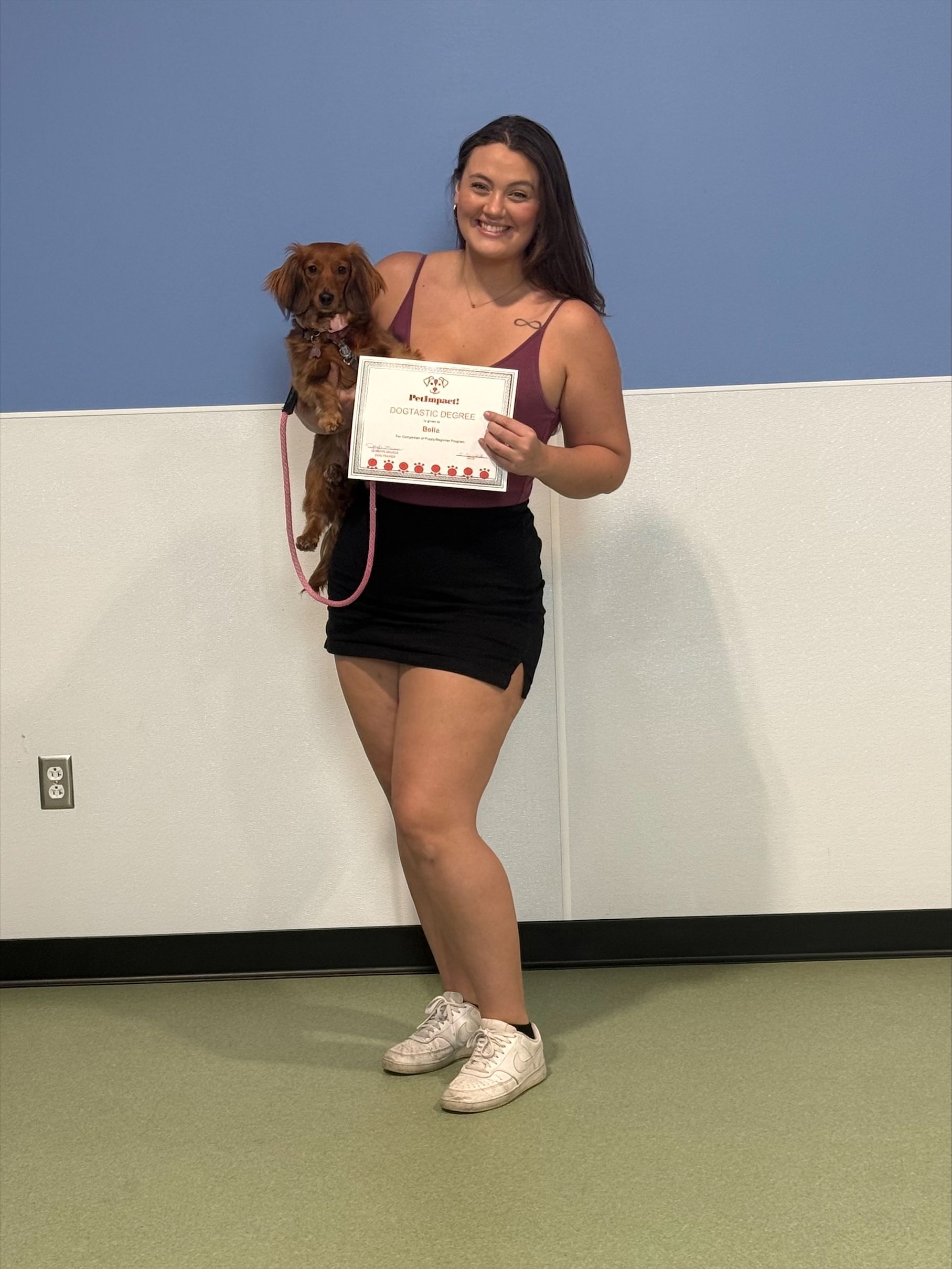 Woman holding a small dog and a certificate, standing against a blue wall and smiling.