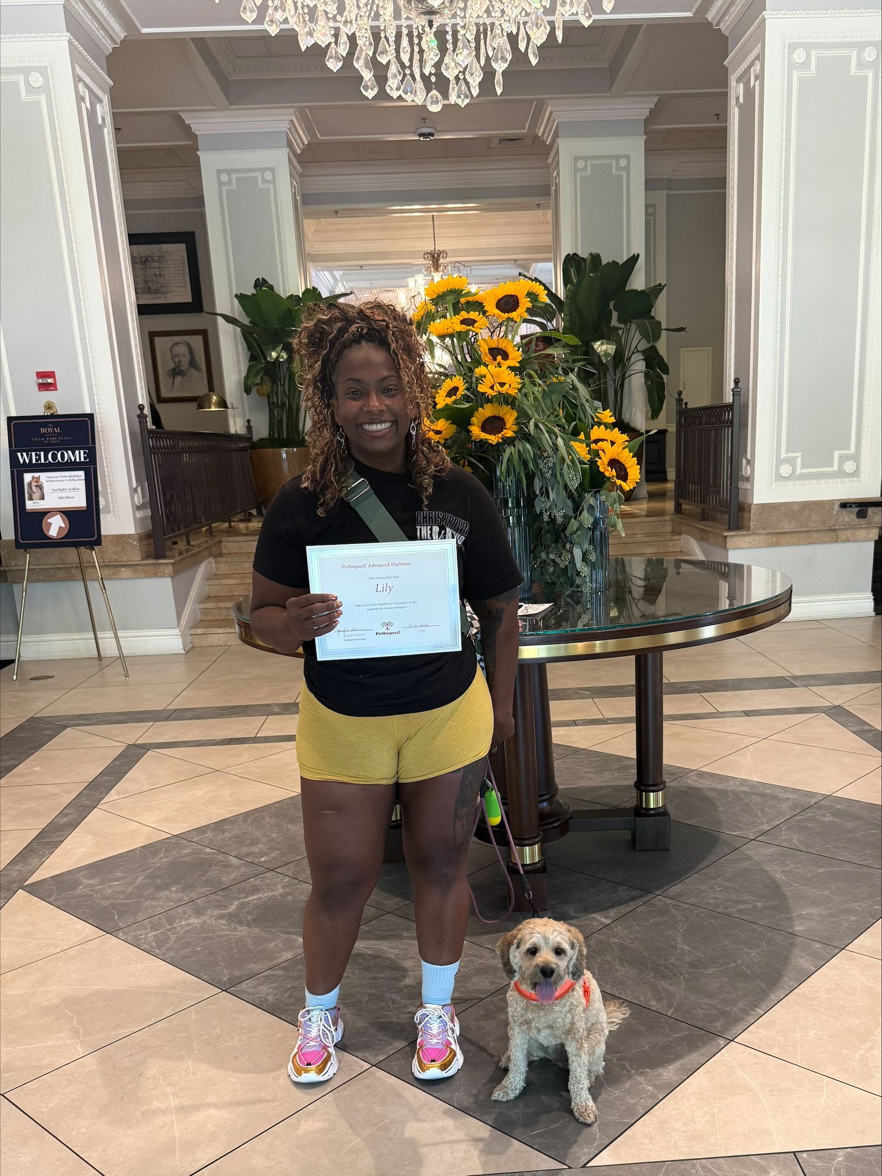 Person holding a certificate with a small dog in a hotel lobby, with a sunflower display behind them