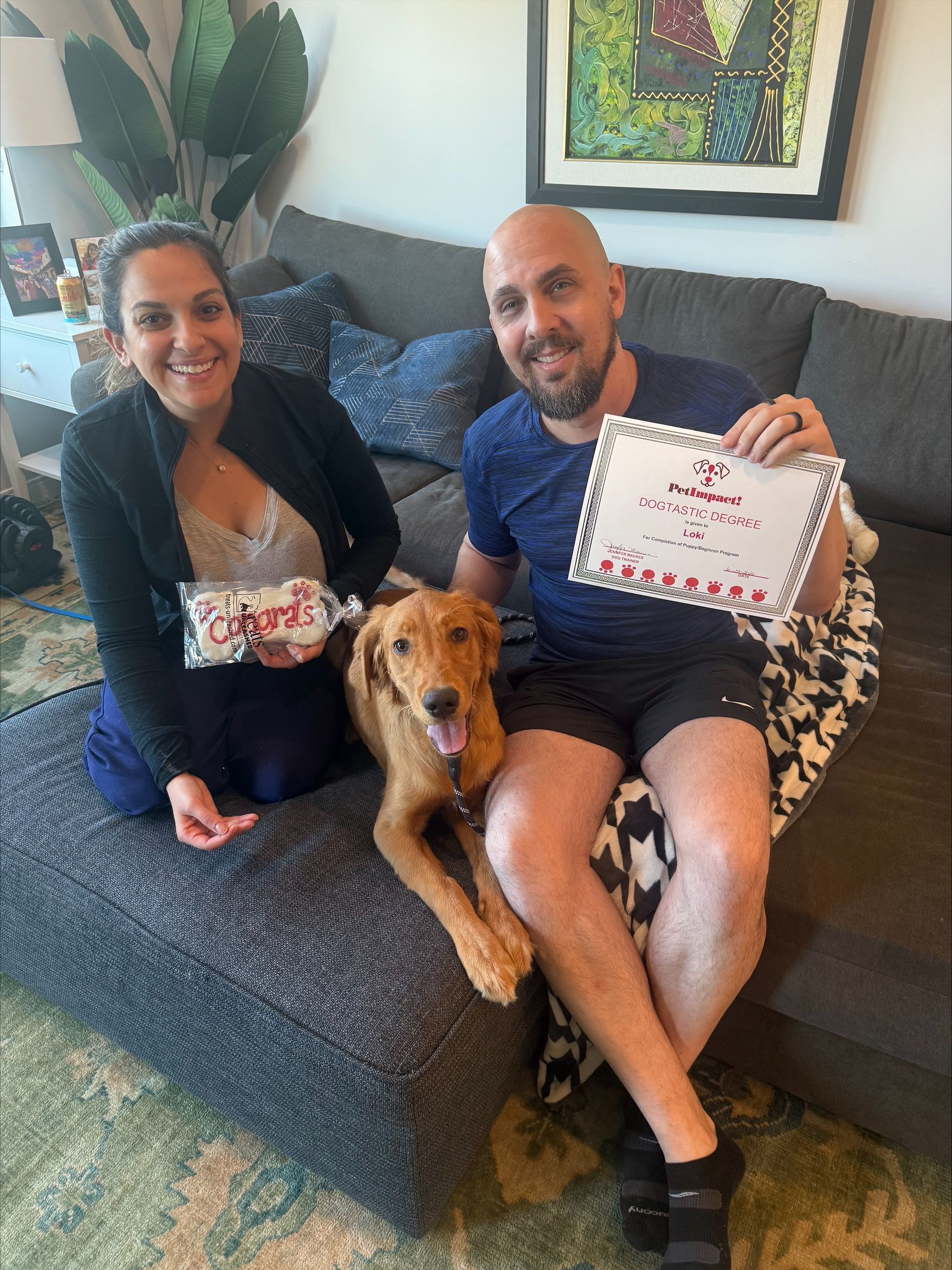 Two people on a couch with a golden dog, holding a certificate and a small floral gift.