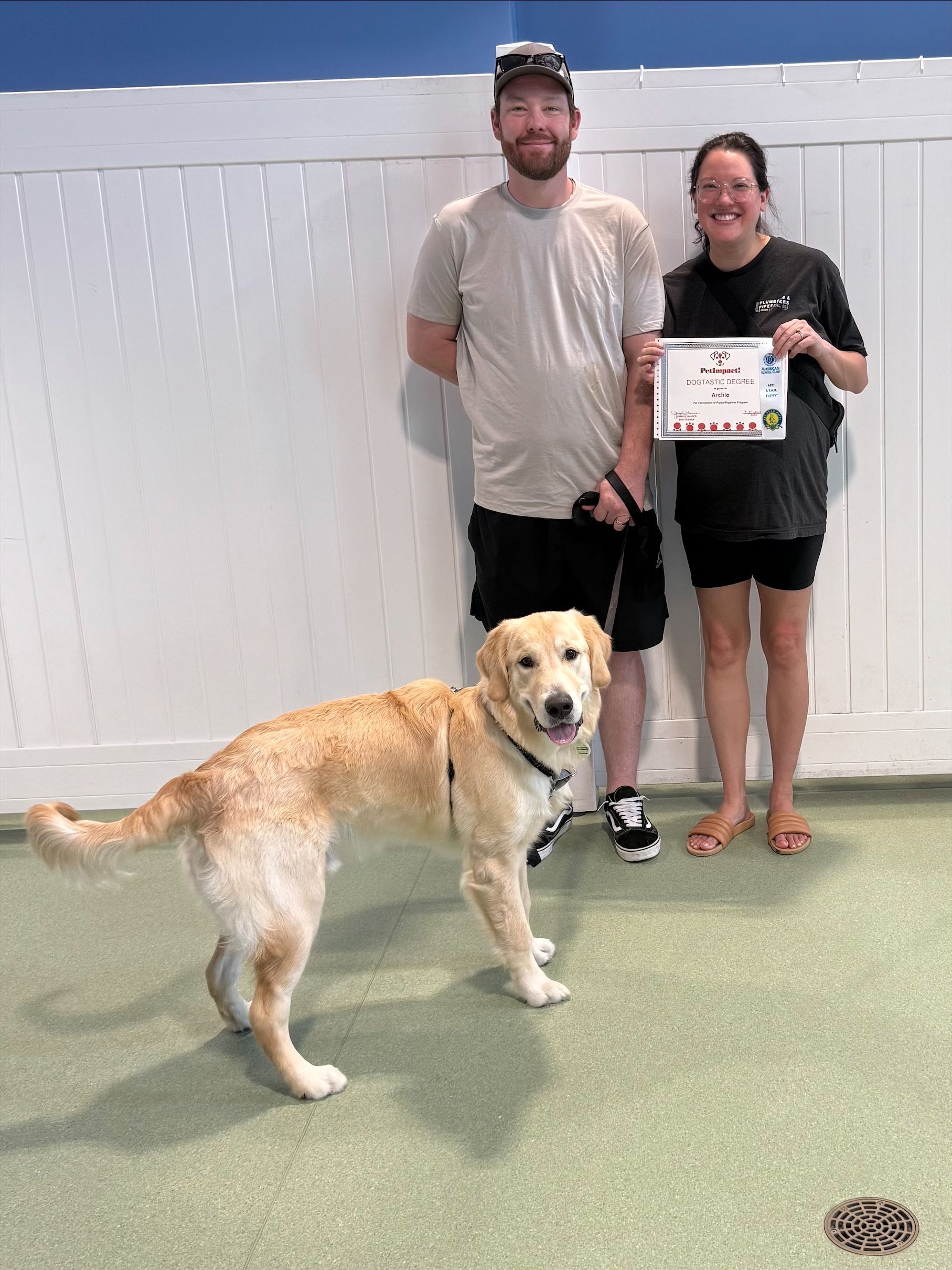 Two people pose with a dog holding a certificate in an indoor room with a white wall.