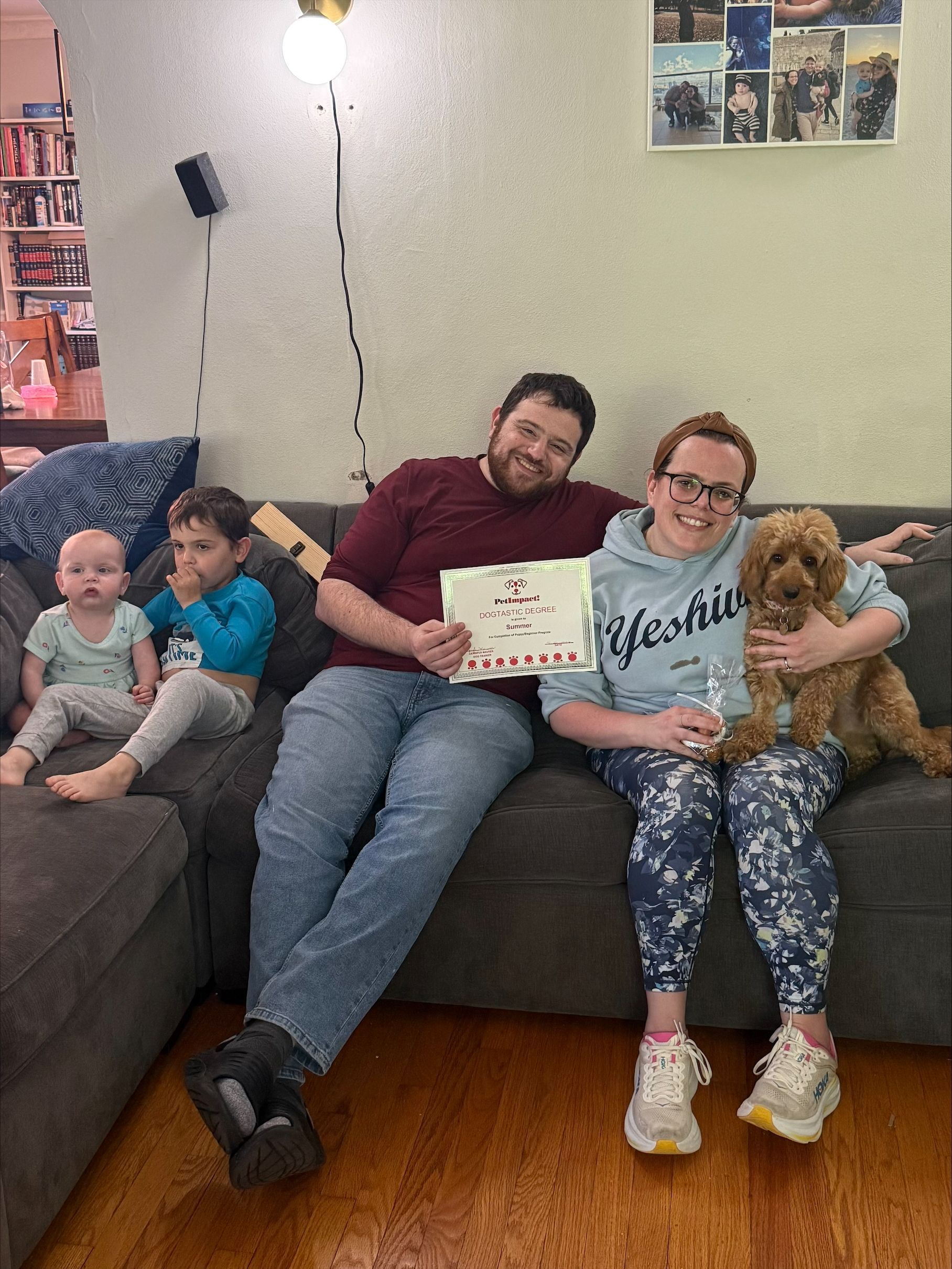 Family sitting on a couch, holding a card and a dog in a living room with a wall light and photo collage.