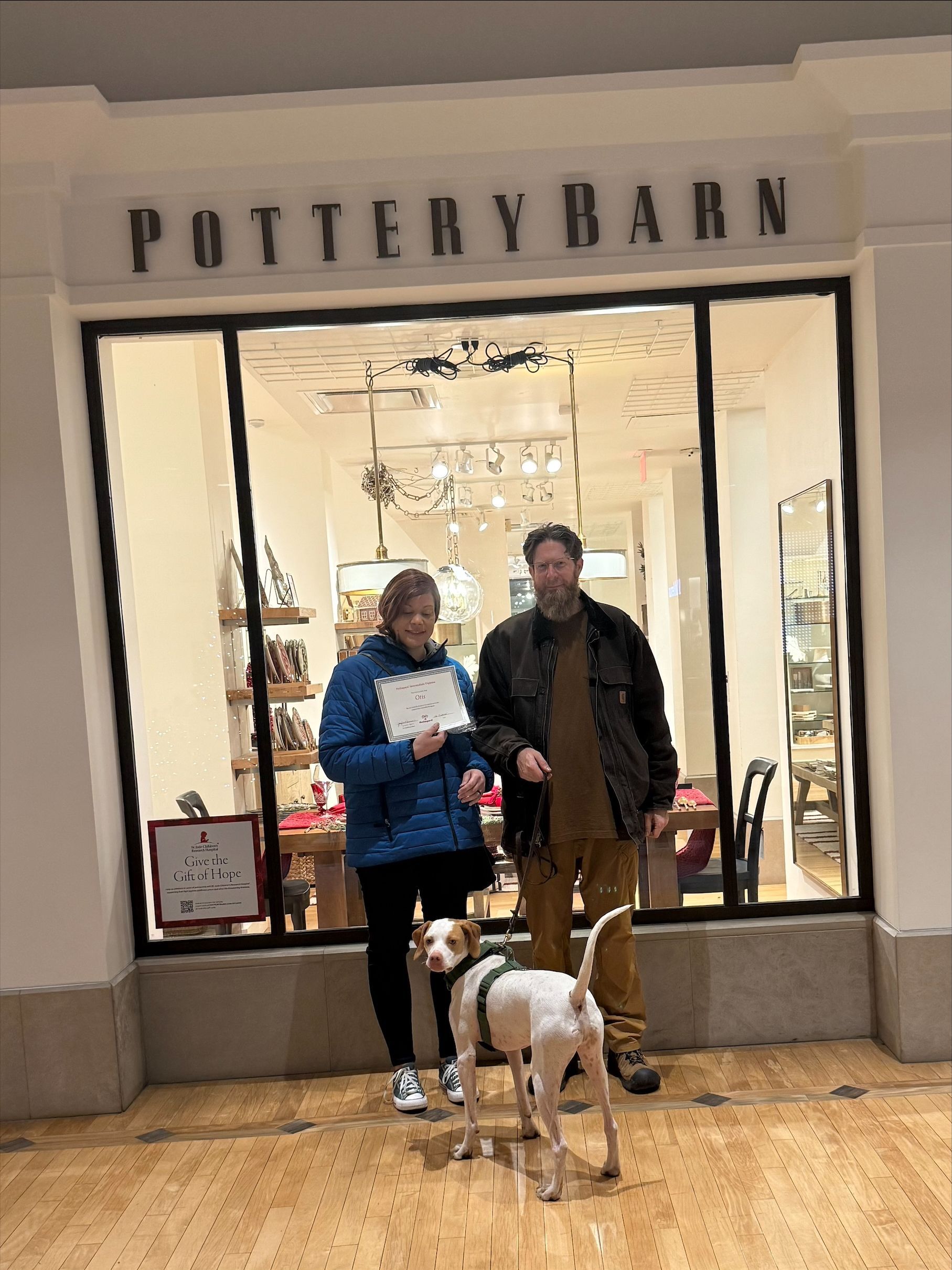 Two people and a white dog stand outside a Pottery Barn store entrance.