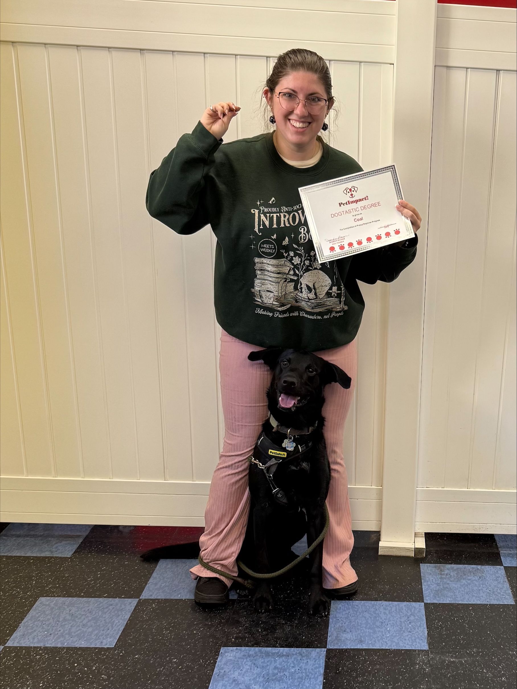 Person with black dog holding a sign, standing indoors and smiling on a checkered floor.