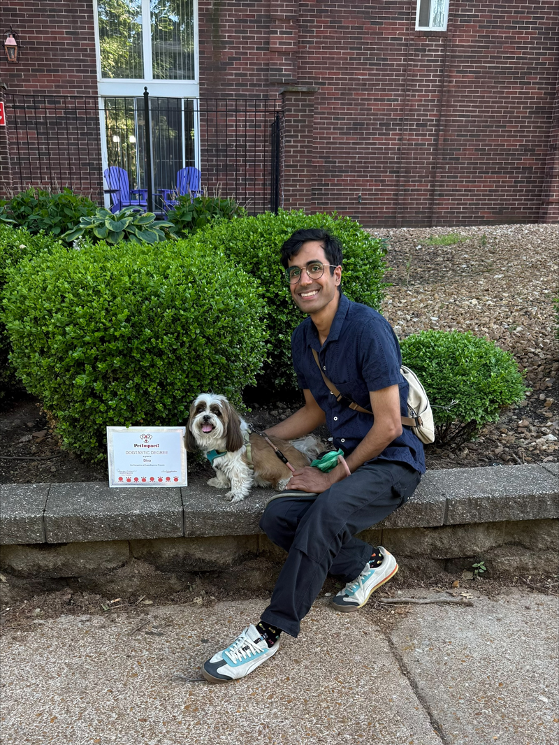 Person sitting on stone steps with a dog beside a small sign, in front of a brick building and bushes.
