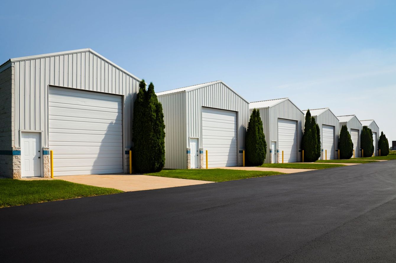 Row of white industrial buildings with garage doors, lined with green shrubbery on a sunny day.
