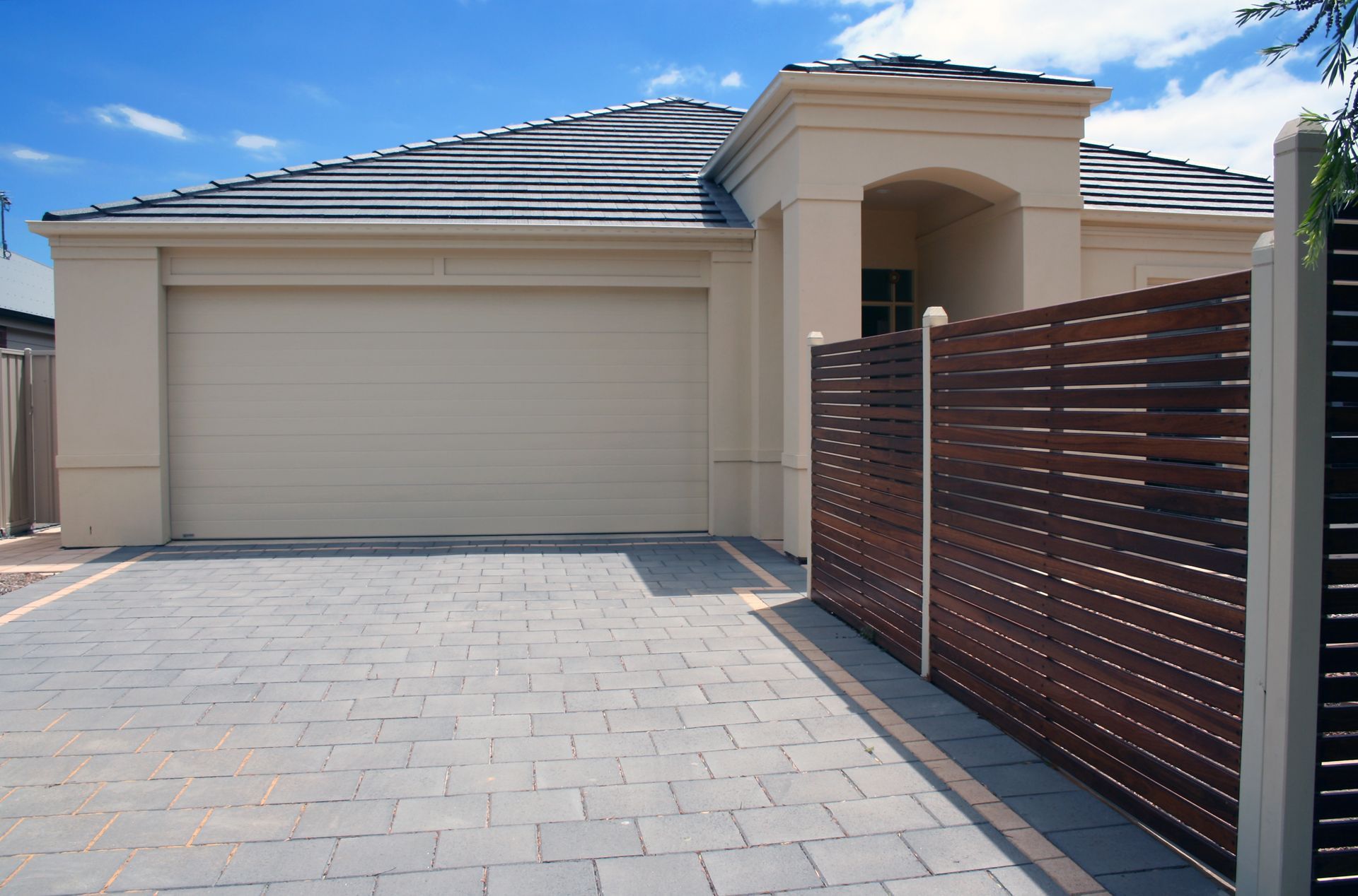 Beige house with a garage, gray paved driveway, and a brown horizontal slat fence under a blue sky.