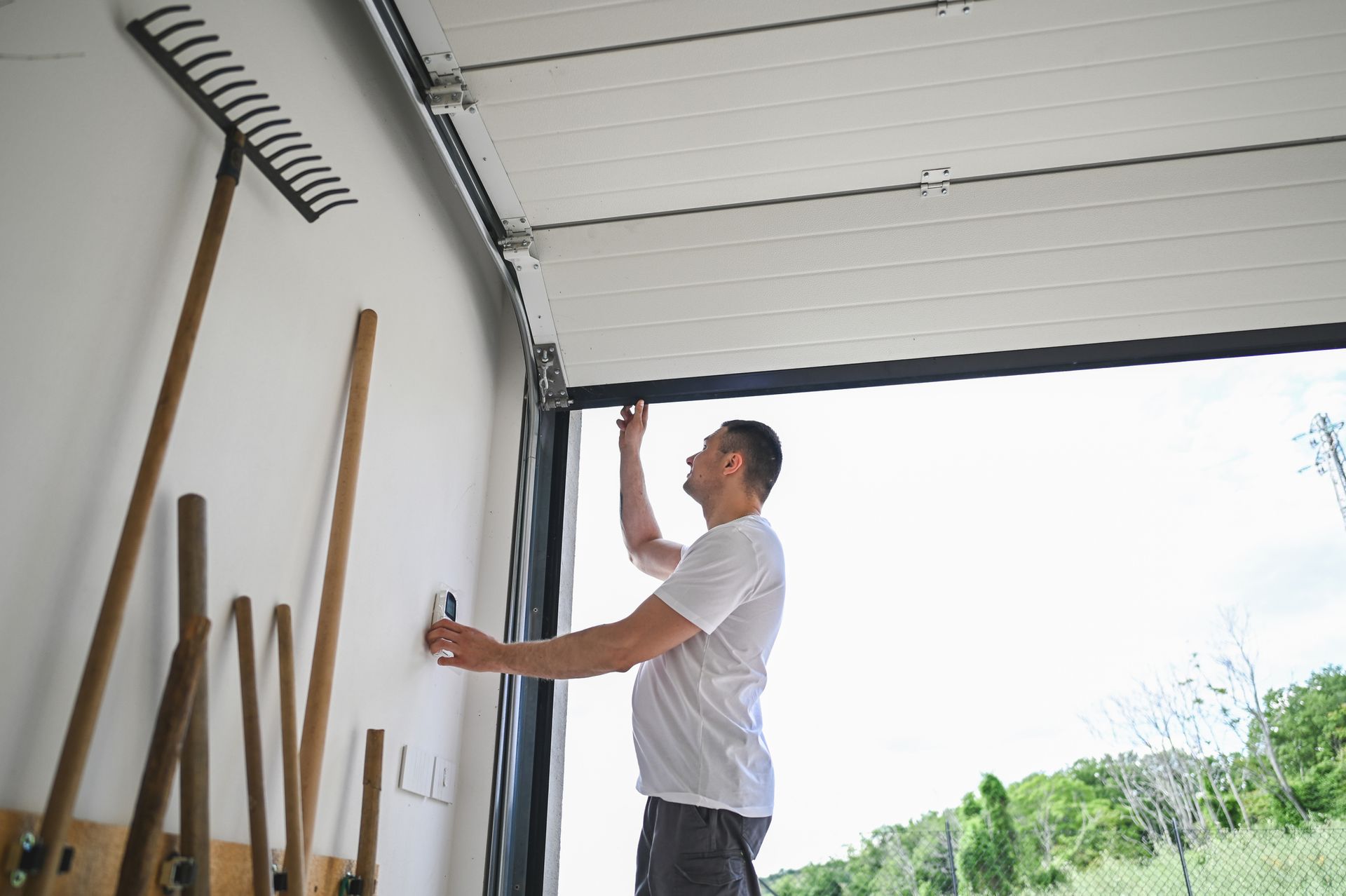 Man inside a garage, reaching up to open the door, tools hanging on the wall.