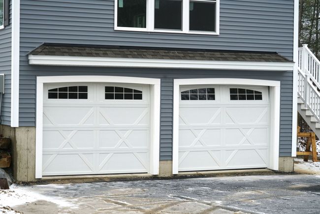 Two white garage doors with windows, in front of a blue house with windows and a dark roof.