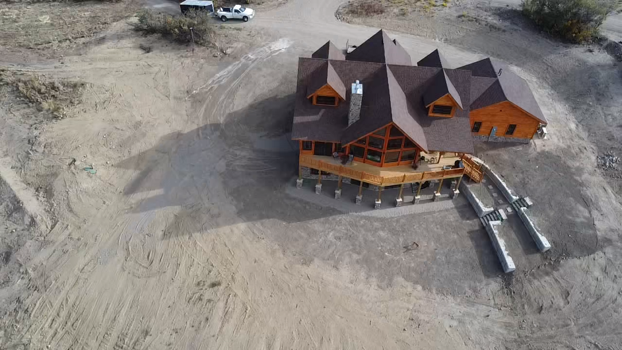 Aerial view of a wooden lodge with dark roofs on a barren, sandy lot and small walkways leading out.