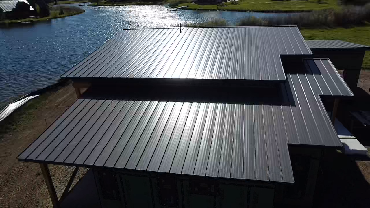 Aerial view of a modern house with a dark metal roof beside a lake.